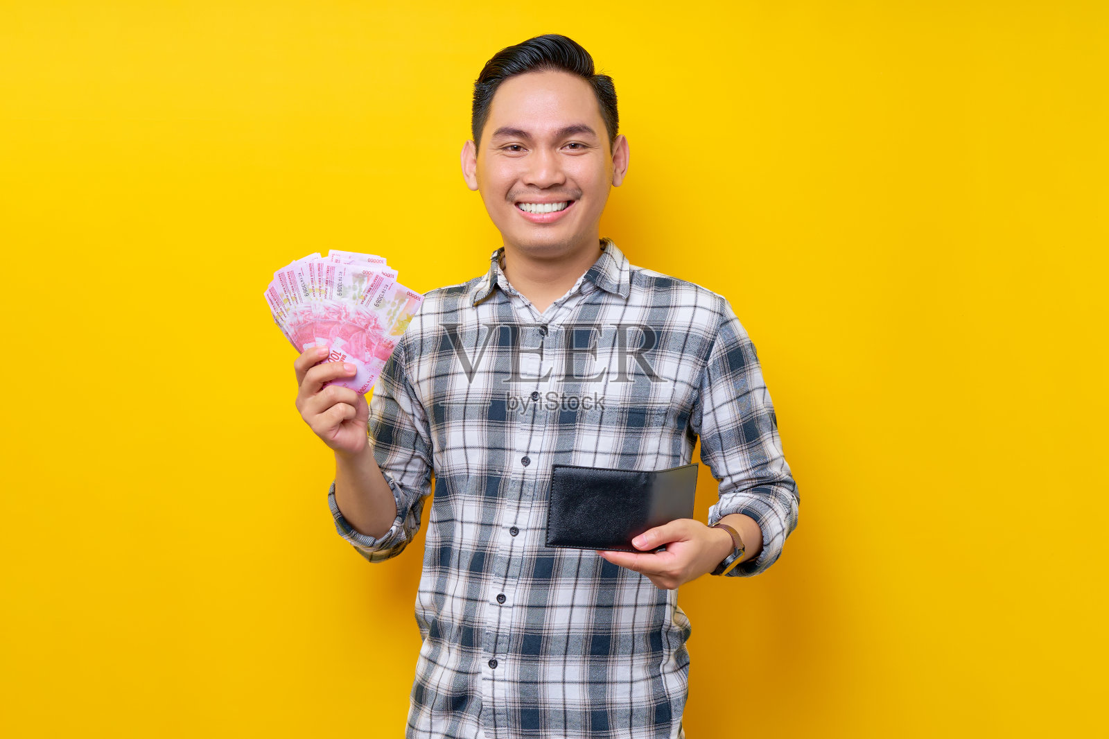 穿着格子衬衫微笑的年轻亚洲男子的肖像，拿着黑色钱包，手里拿着印尼盾纸币，被孤立在黄色背景上。人们的生活方式理念照片摄影图片