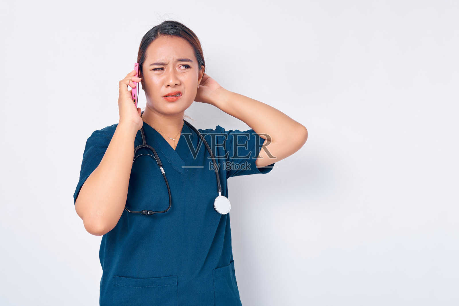 年轻漂亮的亚洲女性，职业护士，穿着蓝色制服，一边讲着手机，在白色的背景上看向一边，脸上露出困惑的表情。保健医学概念照片摄影图片