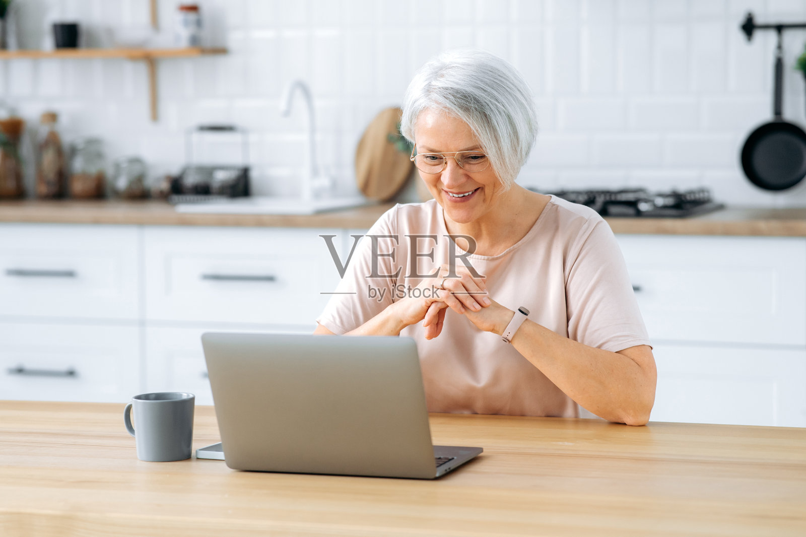 幸福的中年现代白人女士戴着眼镜，坐在家里厨房的桌子旁，拿着笔记本电脑，上网工作，看视频，看新闻，看着笔记本电脑屏幕，微笑着照片摄影图片