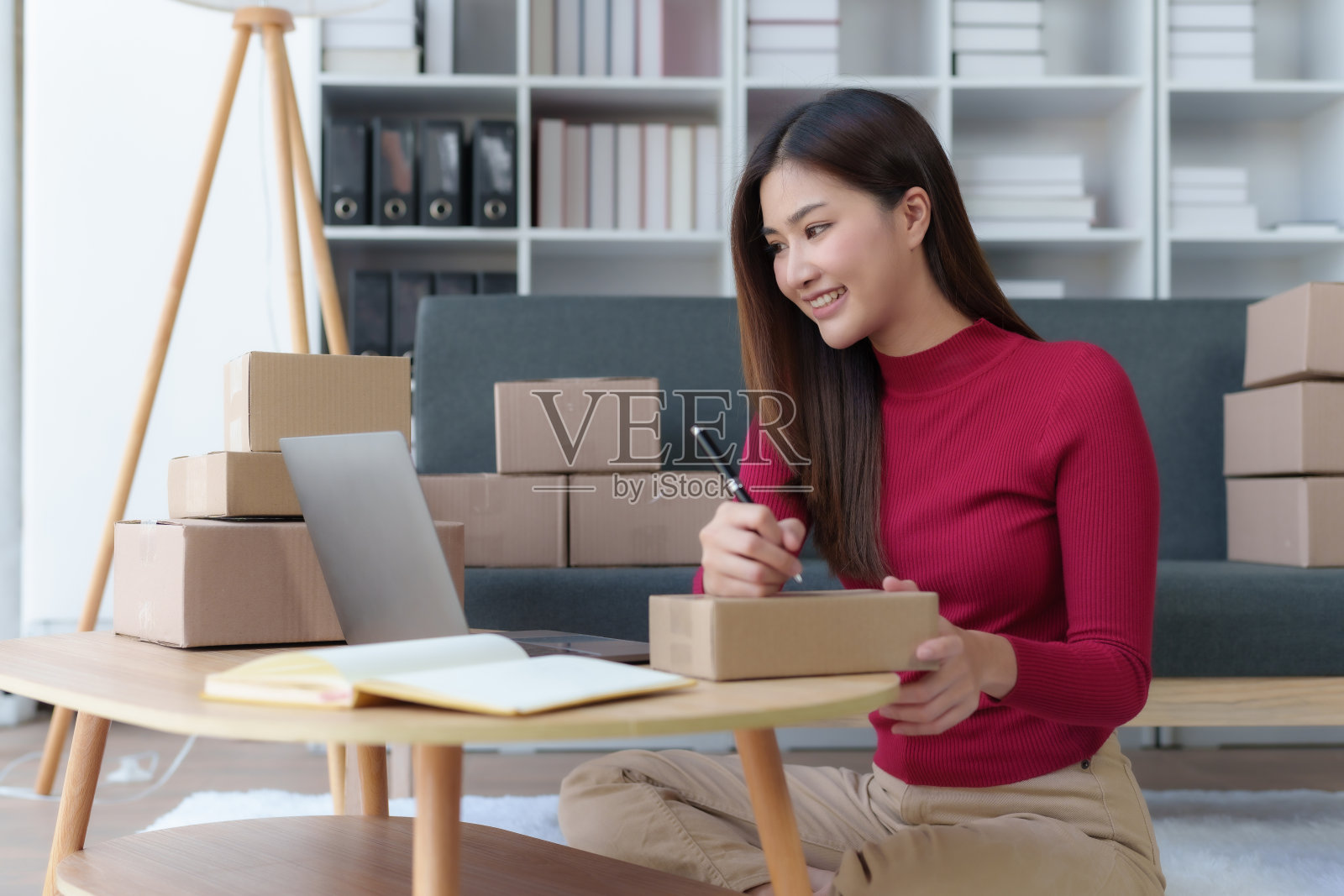 微笑的年轻亚洲女企业主准备包裹箱，在家里的笔记本电脑上检查产品的在线订单。网上购物概念。创办中小企业。照片摄影图片
