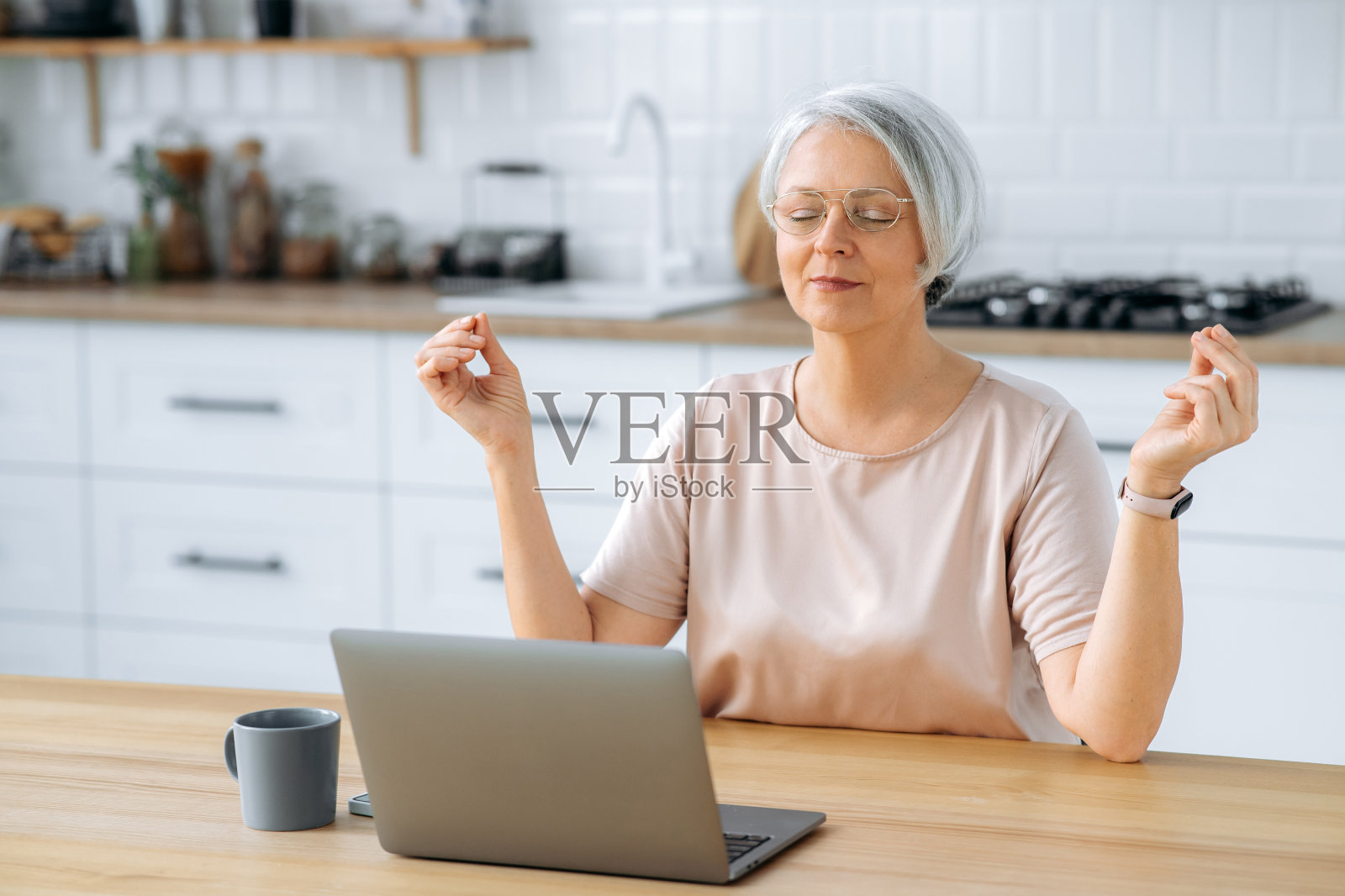 平静、放松、满意的中年白头发、戴着眼镜的白种人妇女，坐在家里厨房的桌子旁，拿着笔记本电脑，在网上工作时放松，闭上眼睛冥想照片摄影图片