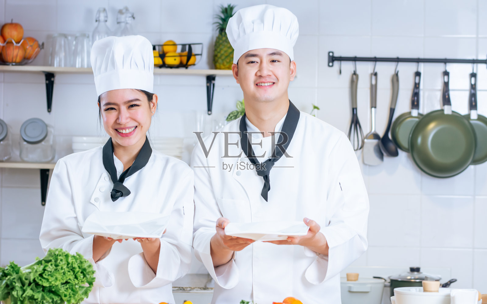 两位亚洲职业厨师夫妇身穿白色制服，头戴帽子，露出空白的盘子，大拇指向上，一起在厨房里做饭，微笑着幸福，充满信心。食品的概念。照片摄影图片