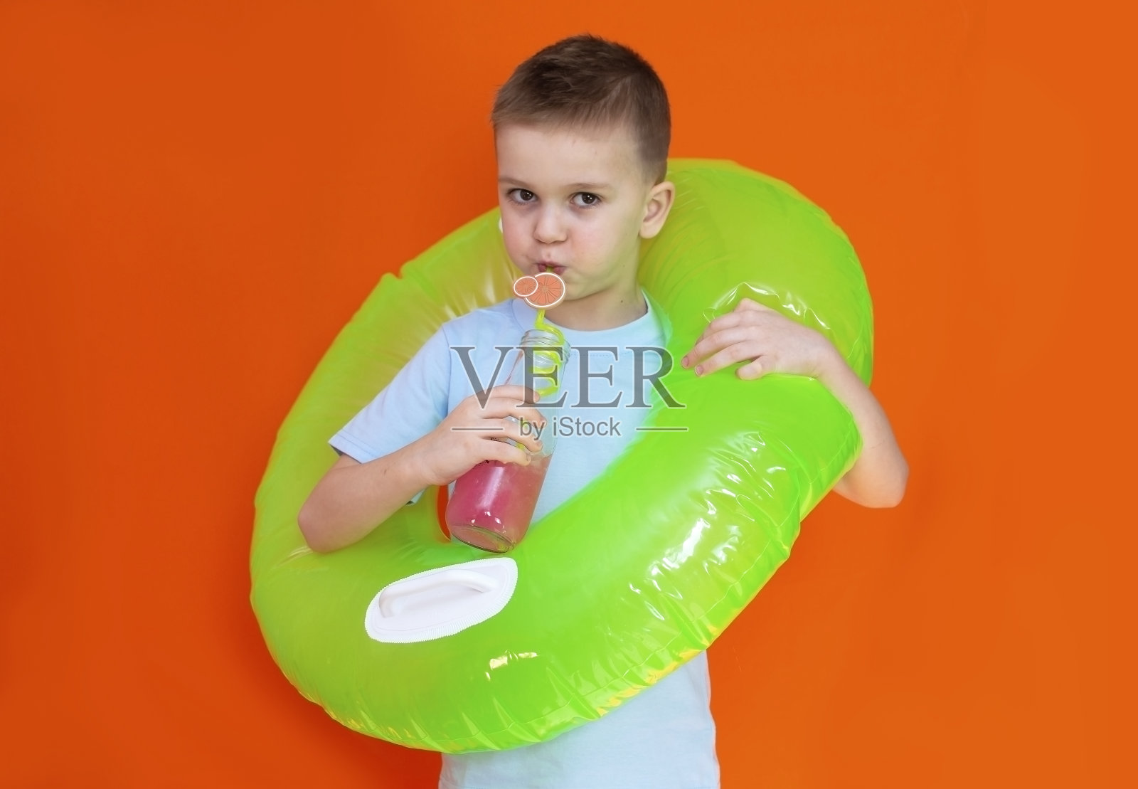 小男孩穿着明亮的t恤，带着一个充气游泳圈，在一个孤立的橙色背景上喝着鸡尾酒。夏日的氛围和休闲的概念。照片摄影图片