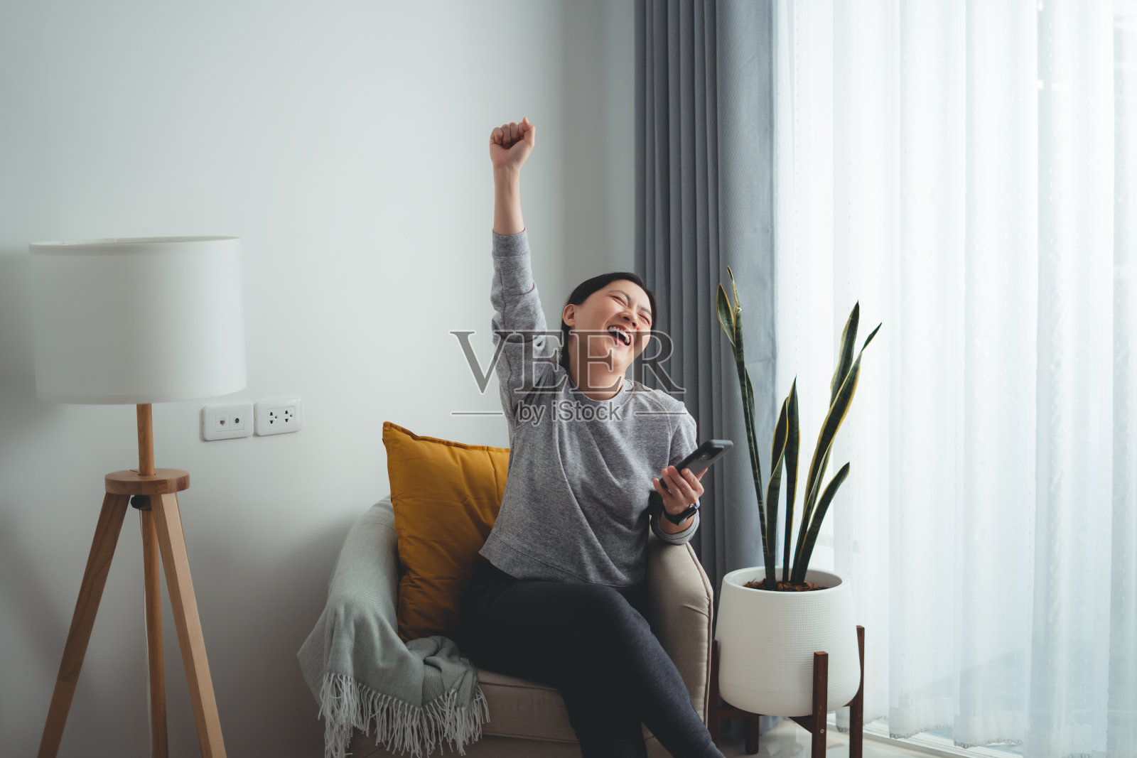 亚洲女性坐在家里的扶手椅上，用智能手机开心兴奋地做出了获胜的手势。照片摄影图片