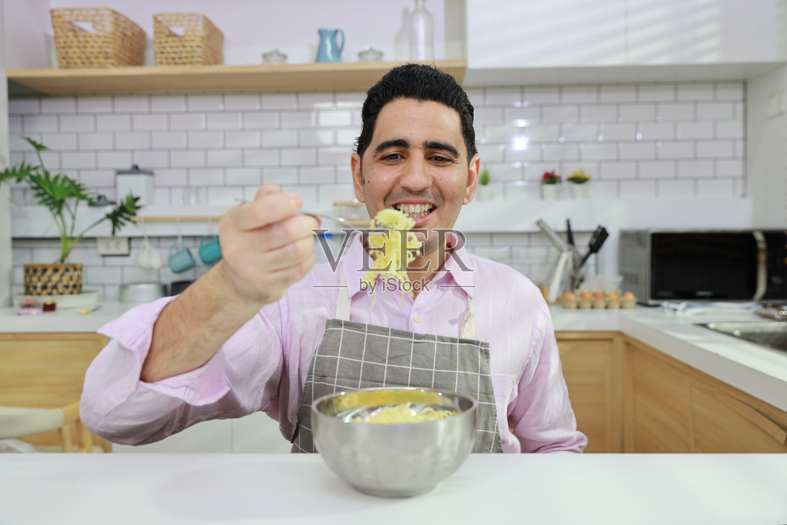 特写年轻快乐的西班牙卷发男人坐在桌子上吃美味的方便面午餐在厨房用筷子和砖墙。不健康和廉价食品的生活方式概念照片摄影图片