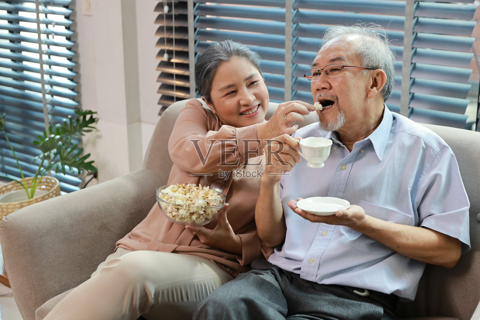 快乐微笑的亚洲老人和女人坐在沙发上吃爆米花，同时在家里的客厅里享受电影休息的乐趣。夫妻俩在看电视时拥抱得很开心照片摄影图片