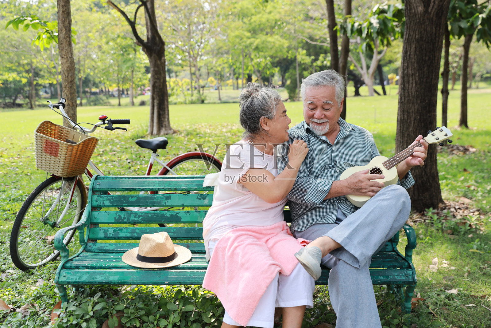 快乐微笑的亚洲老人和女人坐在长凳上弹尤克里里和唱歌在花园公园户外。音乐和放松使情侣幸福。养生生活理念。照片摄影图片