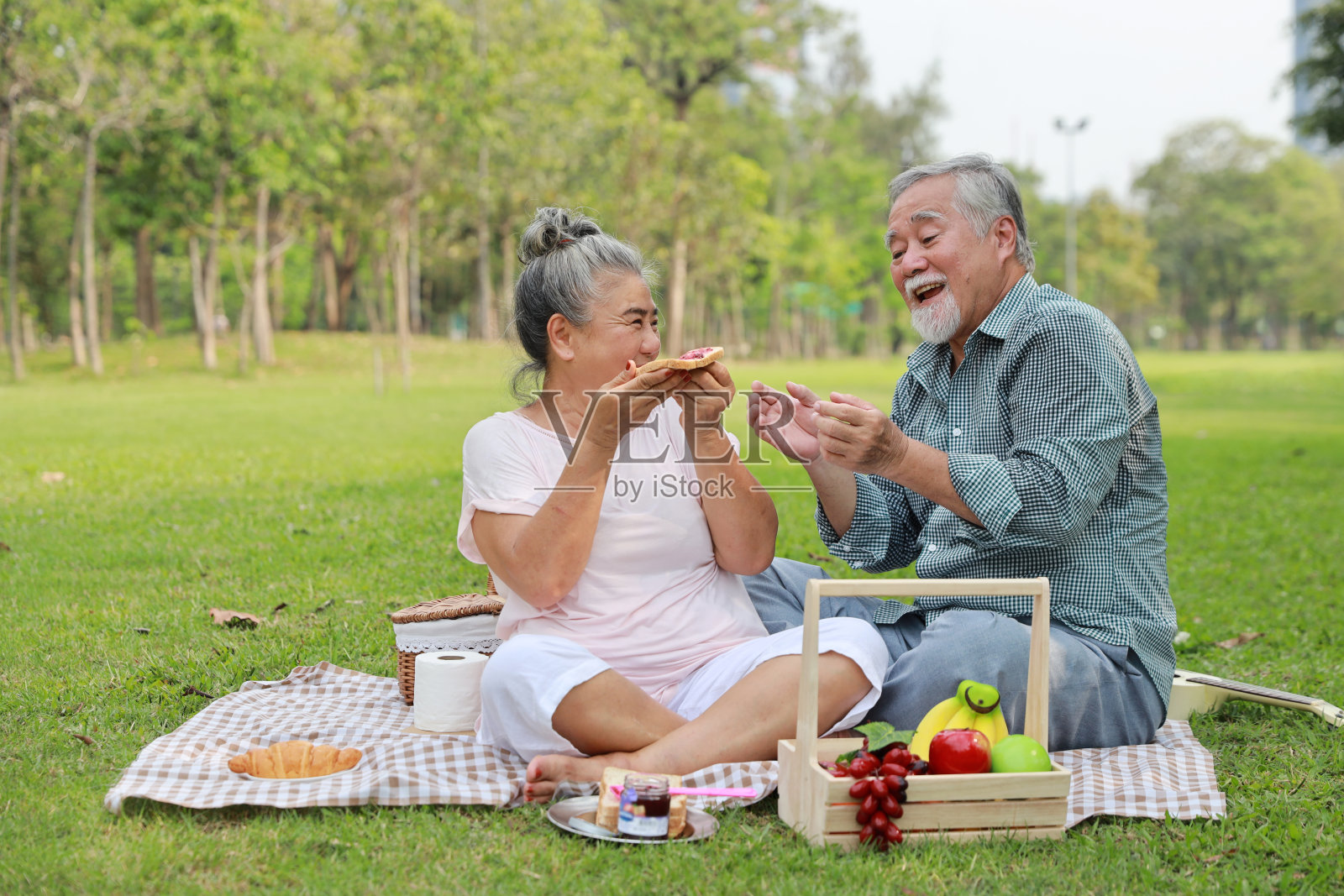 快乐的亚洲老人和女人坐在毯子上，在户外花园一起野餐。情侣们在公园里吃着食物，拥抱在一起。幸福婚姻生活理念。照片摄影图片