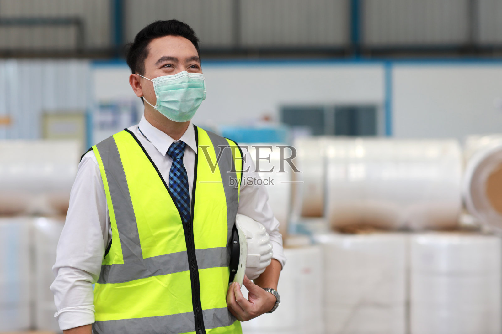 亚洲工程工人肖像站着，拿着安全帽，戴着防护面具，在模糊的大仓库背景下充满自信照片摄影图片