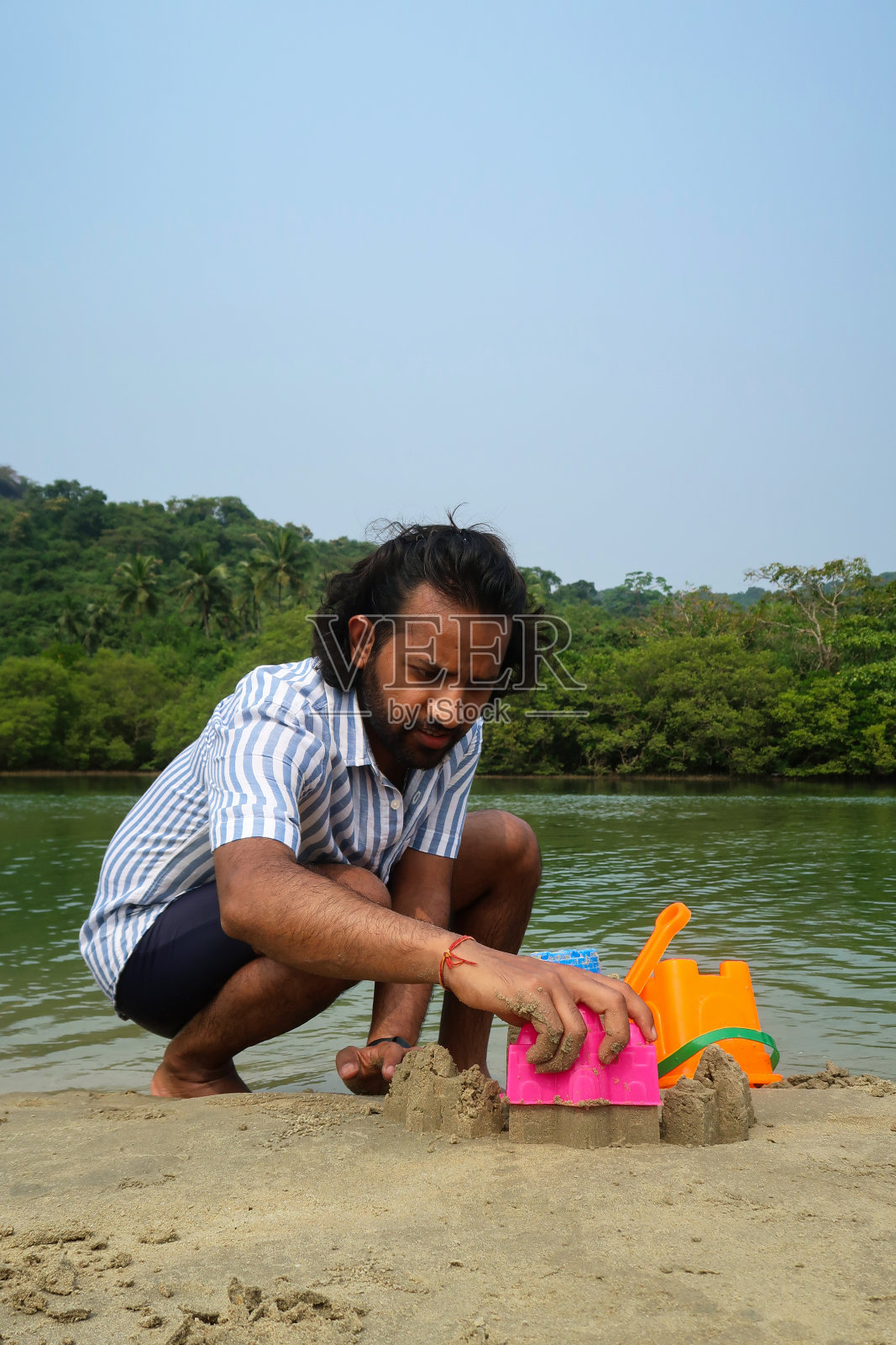 印度男子穿着冲浪短裤和条纹短袖衬衫蹲在泻湖水边的沙滩上，用塑料堆沙堡，色彩鲜艳的儿童海滩玩具，水桶和铁锹，看到长满棕榈树的林地照片摄影图片