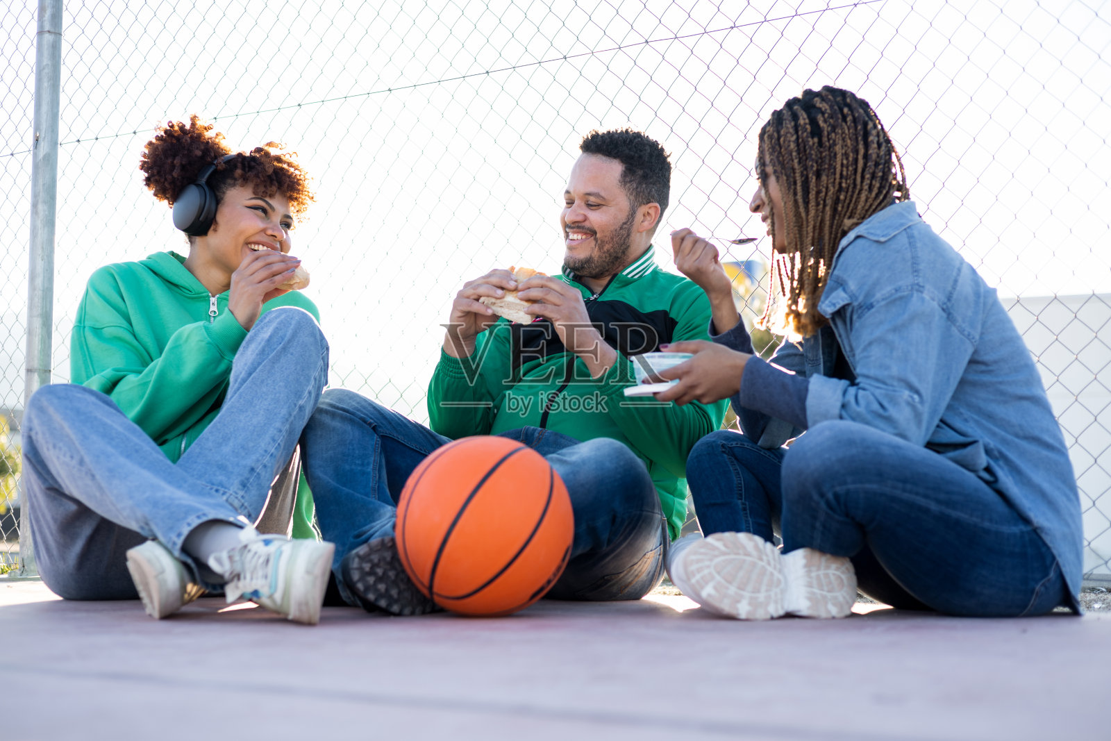 一群快乐而多样的朋友聚集在篮球场上。三个朋友坐在一起吃东西。不同种族的人之间的社会关系概念。照片摄影图片