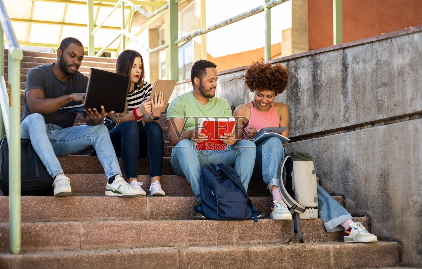 一群穿着随意的大学生。4个年轻人坐在大学的楼梯上，拿着课本、笔记本电脑和平板电脑学习。千禧一代大学生的概念。照片摄影图片