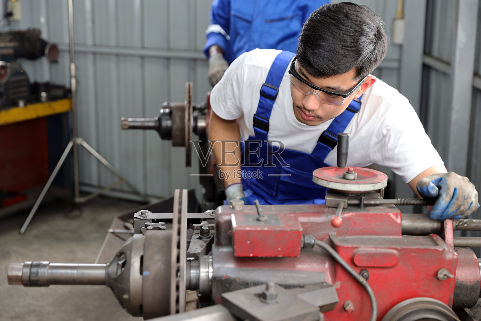 身穿防护服的多民族技工或工人在车库用金属车床抛光汽车盘式制动器。维护汽车和检查汽车零件的概念照片摄影图片