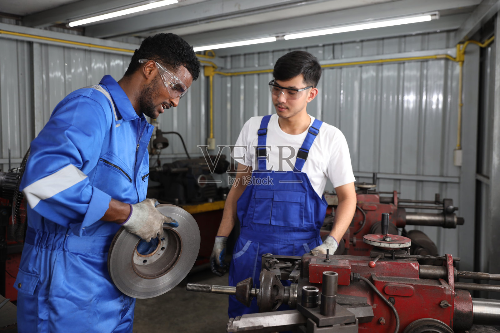 身穿防护服的多民族技工或工人在车库用金属车床抛光汽车盘式制动器。维护汽车和检查汽车零件的概念照片摄影图片