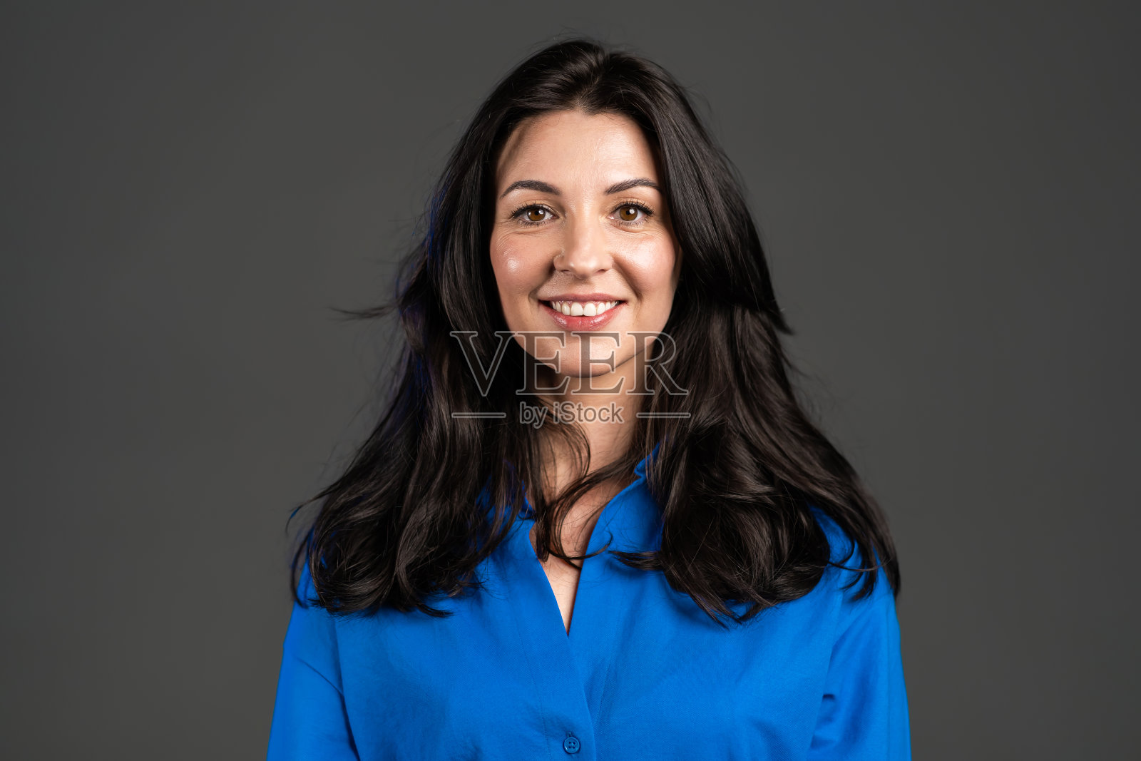 迷人的成熟女子孤立在灰色的工作室背景。成年女士的肖像，长头发，穿着蓝色衬衫，微笑着看着镜头。照片摄影图片