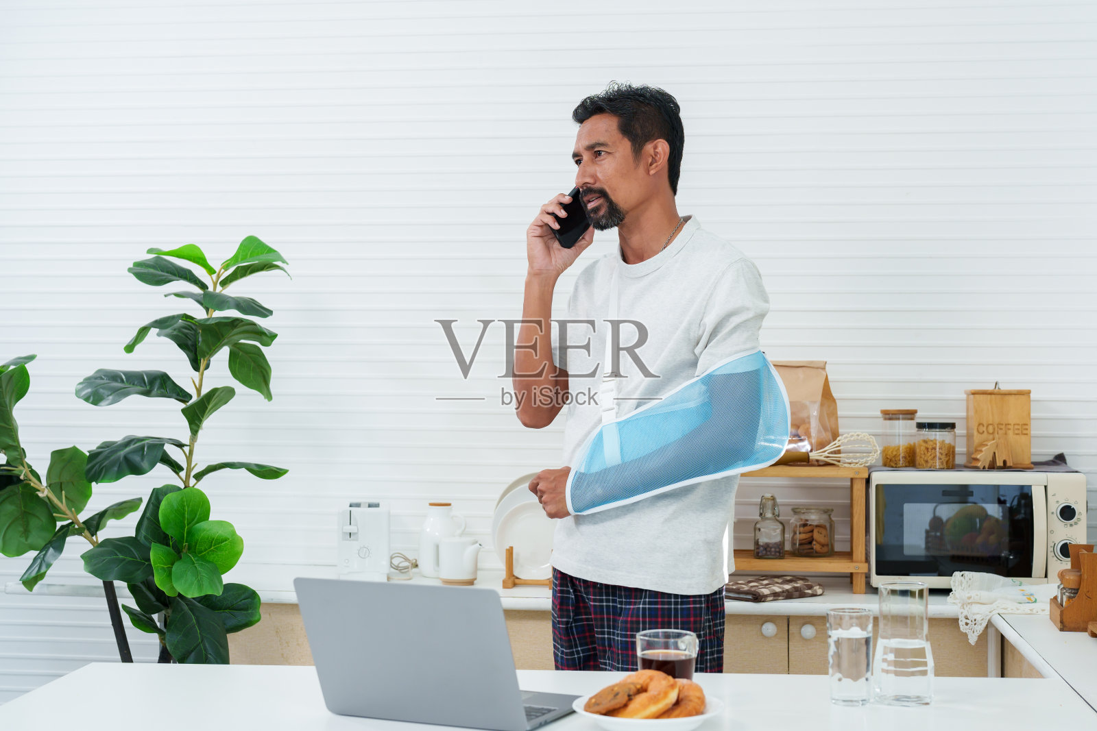 一名亚洲男子左臂受伤，需要康复治疗后在家工作。使用恢复臂支架。用智能手机咨询同事。通过笔记本电脑在线工作照片摄影图片