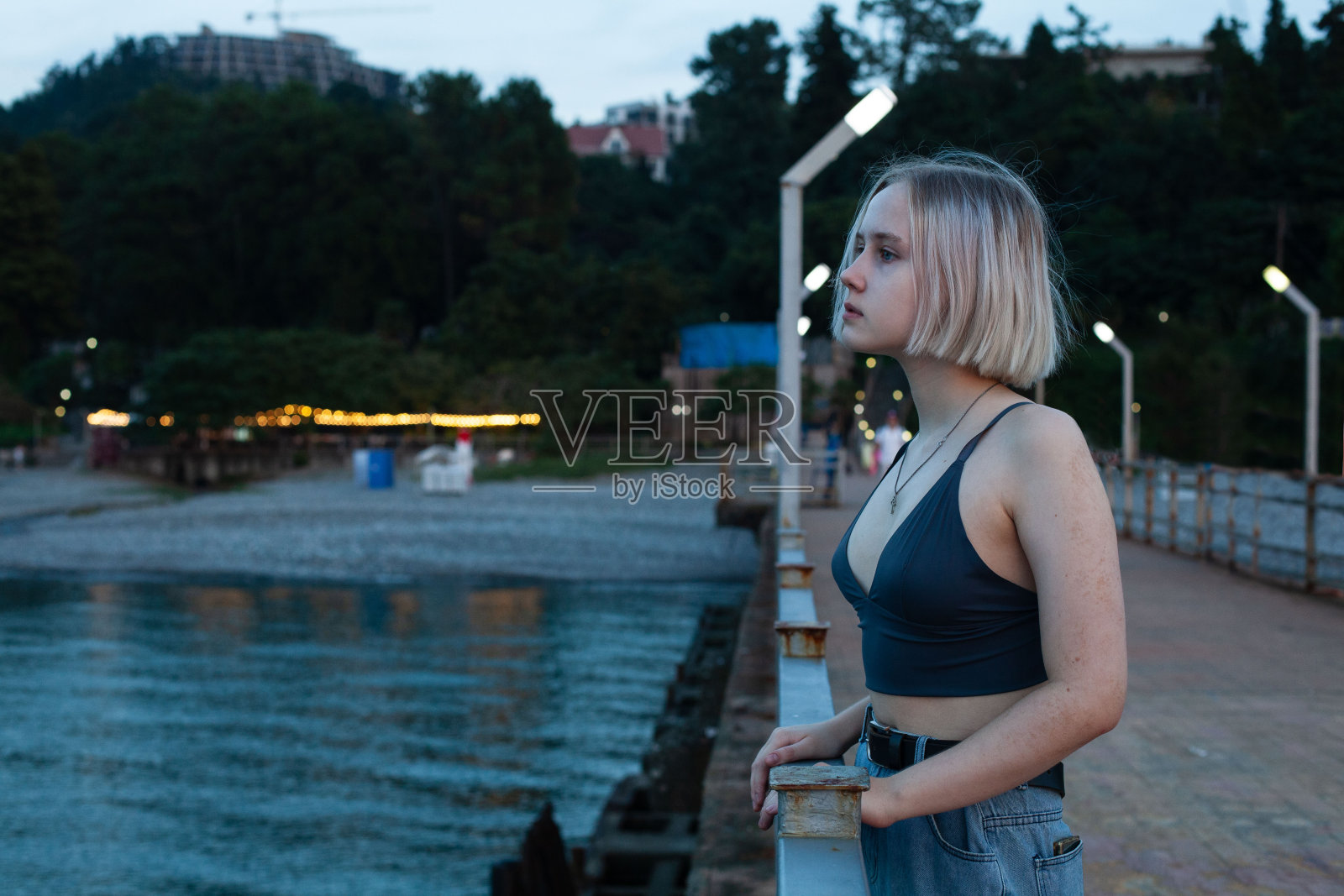 身穿背心的年轻金发女子站在码头上，背景是蓝色的大海和傍晚的海滩公园照片摄影图片