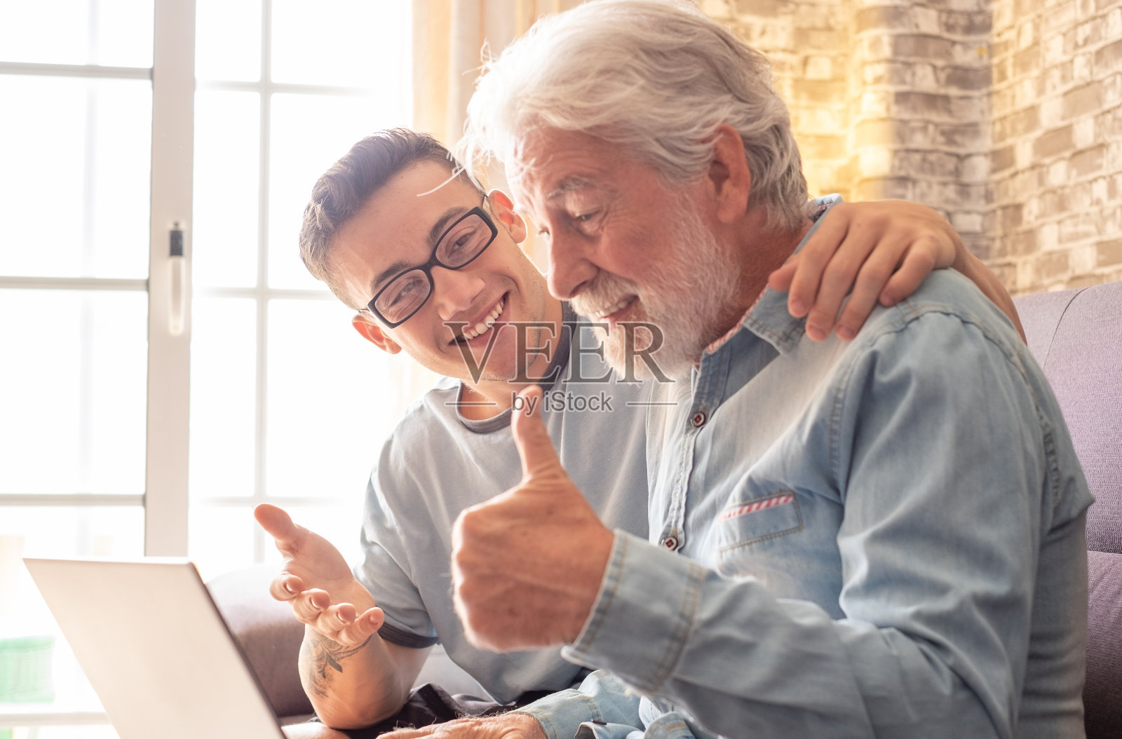 老人和小男孩坐在家里的沙发上一起看笔记本电脑。侄子教并展示新的电脑技术电子商务网上购物给他的祖父。照片摄影图片