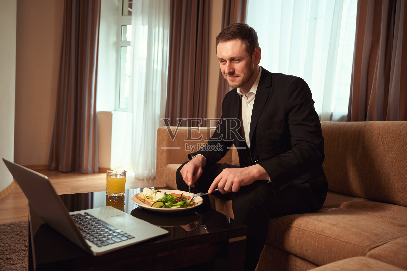 微笑的男人在桌子边吃饭，看着笔记本电脑照片摄影图片