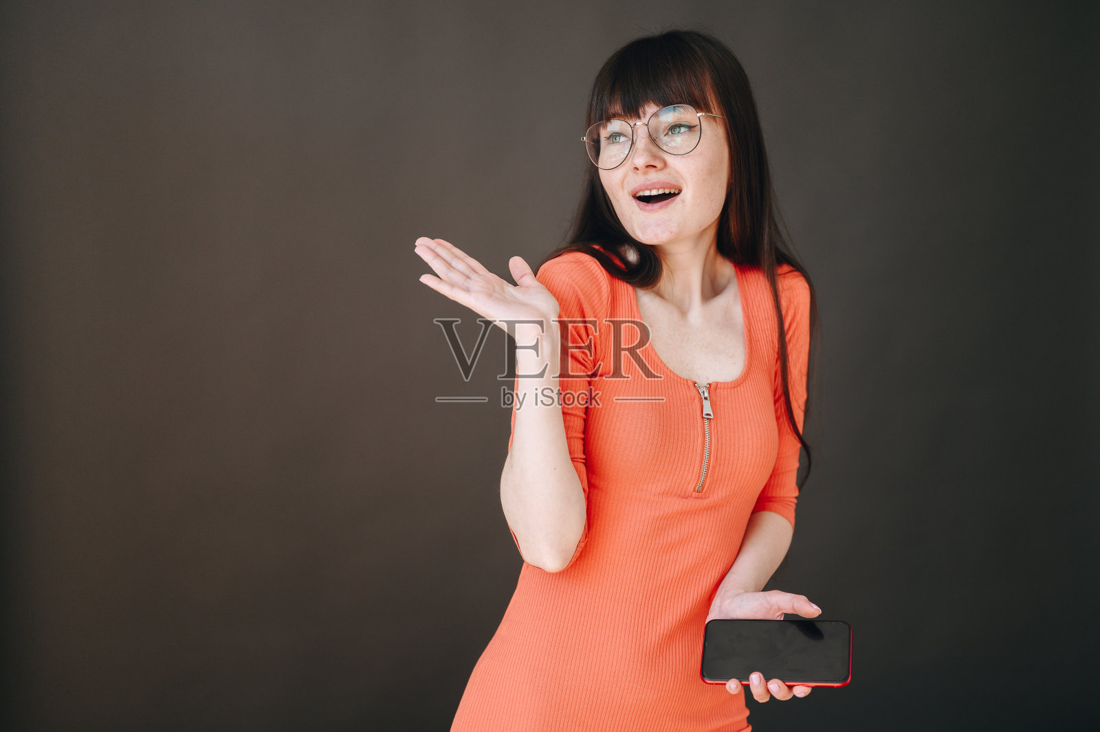 一个戴眼镜的年轻女孩拿着手机，在黑色背景上情绪激动地摆姿势照片摄影图片