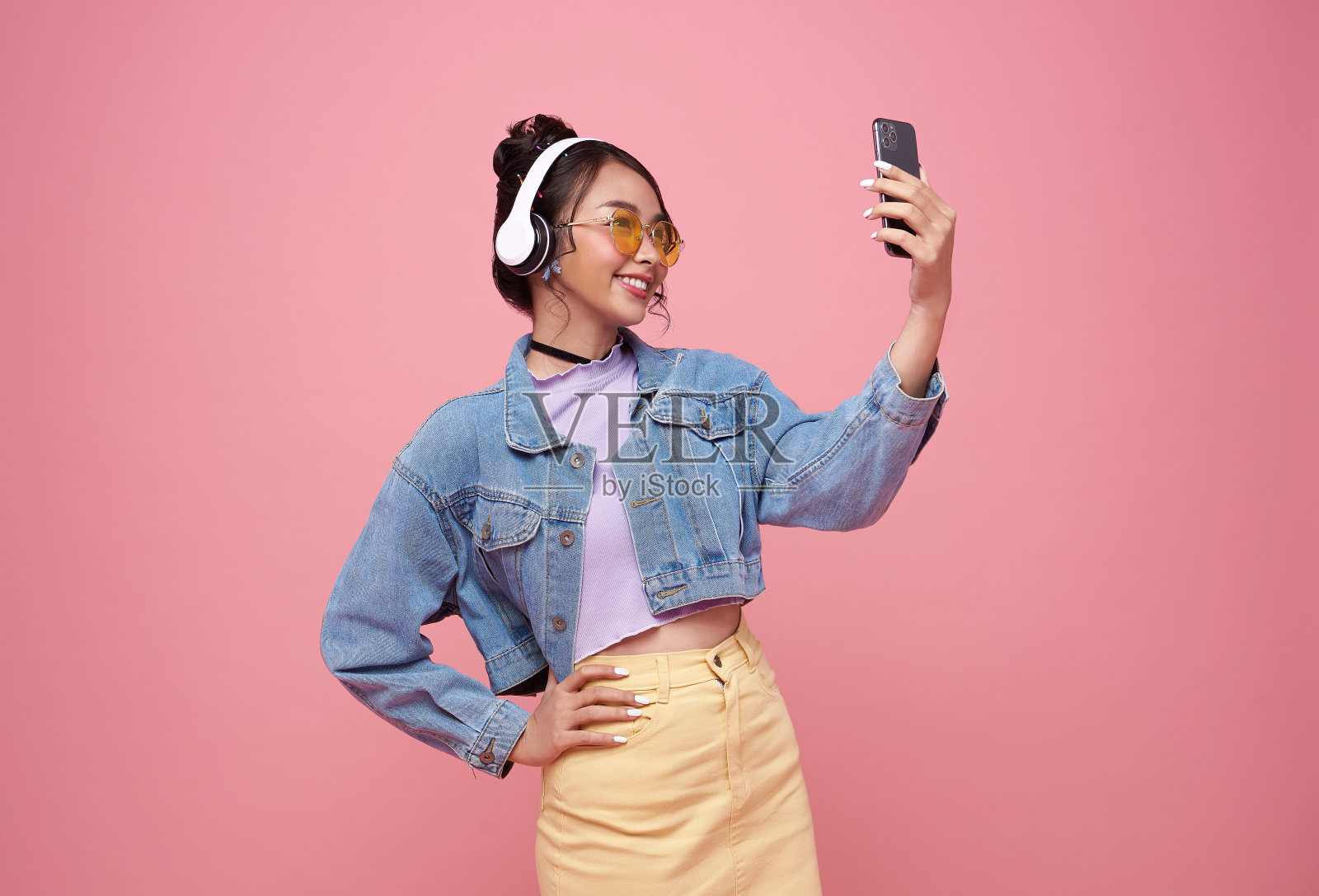 年轻的亚洲少女使用智能手机，她戴着耳机听着粉红色背景音乐。照片摄影图片