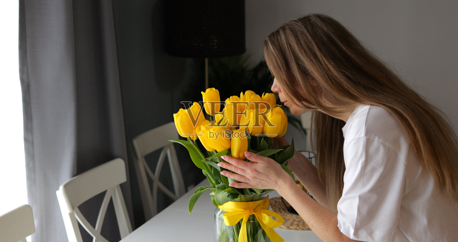年轻女子捧着花束倾身闻着春天黄色荷兰郁金香的香气。装饰玻璃花瓶营造温馨的节日气氛。照片摄影图片