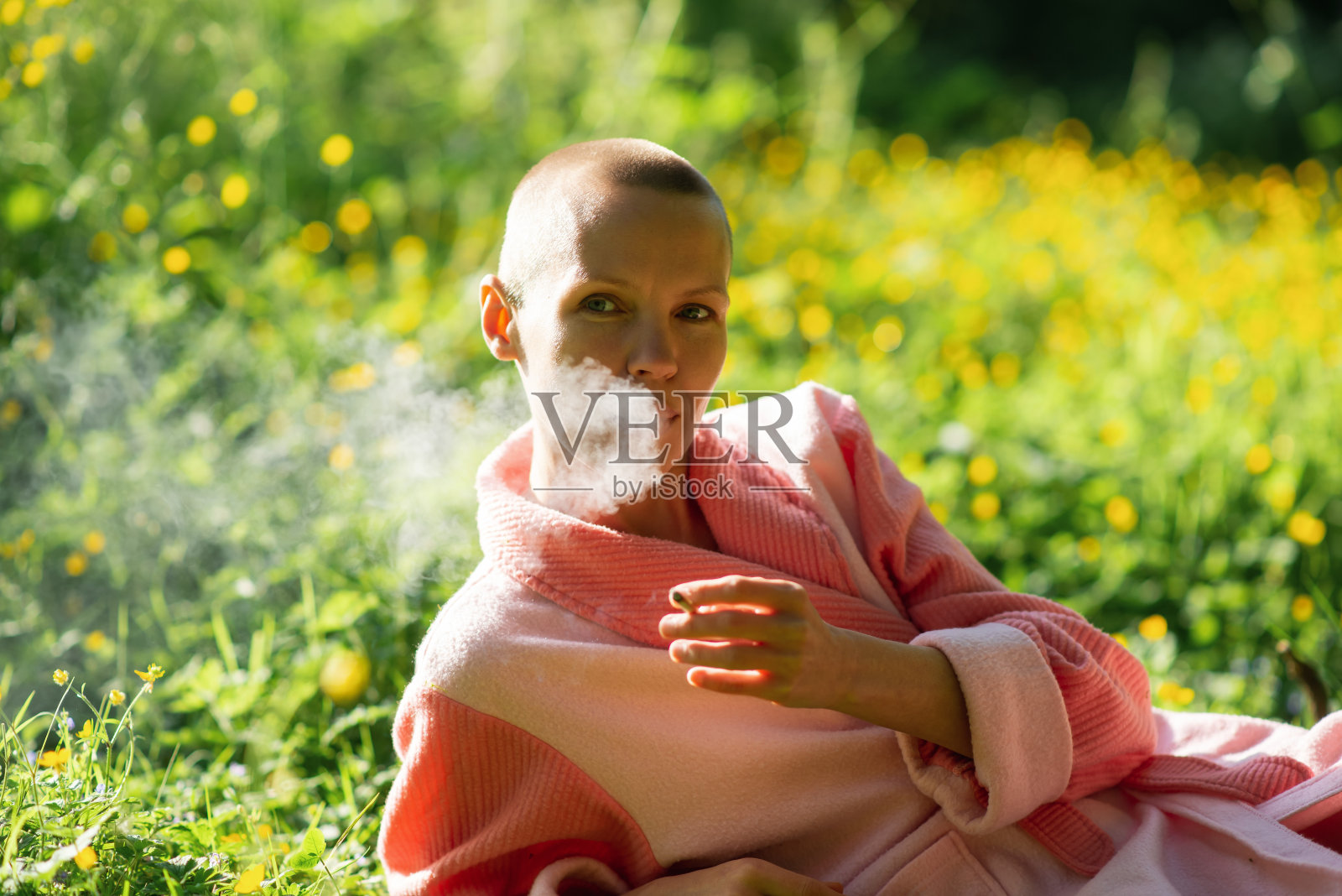 一个没有头发的瘦弱女人躺在绿草地上抽烟。化疗的结果，恢复。照片摄影图片