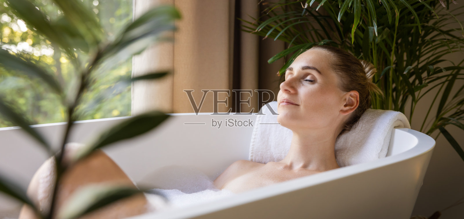 在有绿色热带植物的浴室里，女人闭着眼睛在浴缸里放松。水疗、身体及皮肤护理。带有复制空间的横幅照片摄影图片
