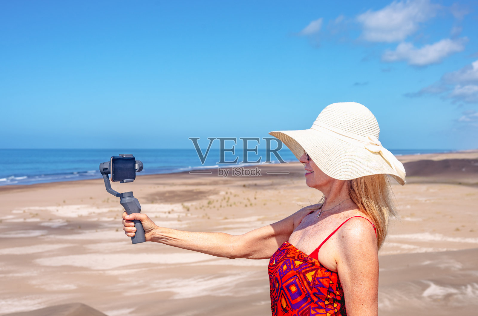 一个成熟的女人在沙滩上的三脚架上用手机自拍。照片摄影图片