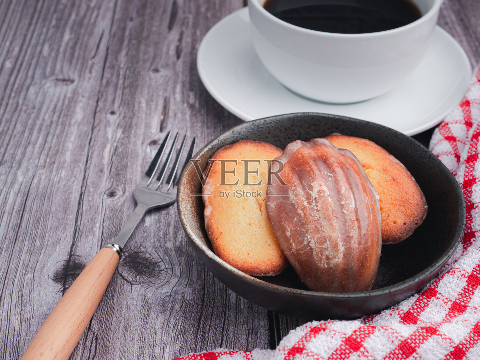 玛德琳自制的传统法式小饼干放在黑色的盘子上，用一块布和一个咖啡杯放在木桌上。照片摄影图片