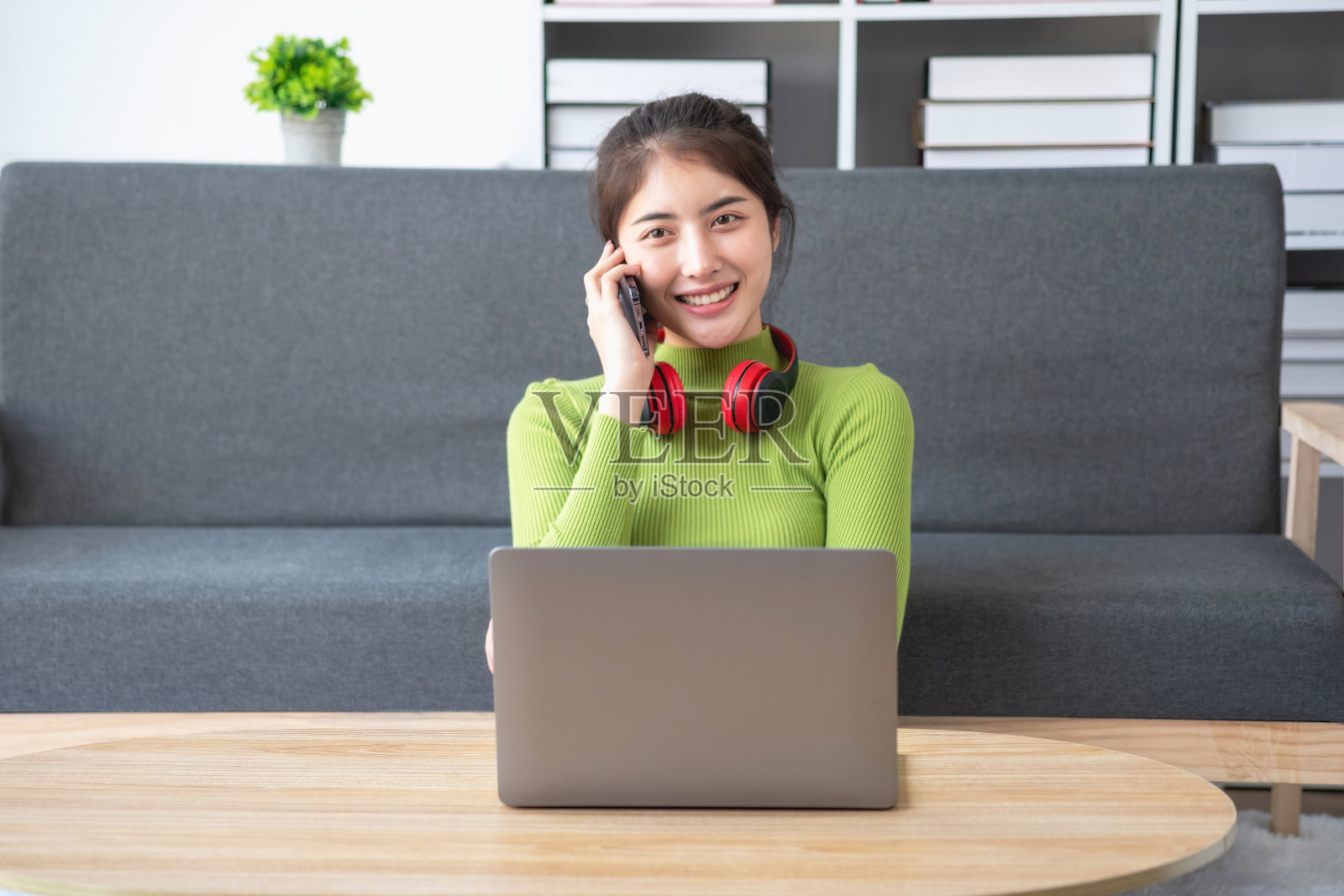 亚洲美女穿着绿色长袖衬衫，悠闲地坐在家里的客厅里，用笔记本电脑、手机和耳机看电影，听音乐。照片摄影图片