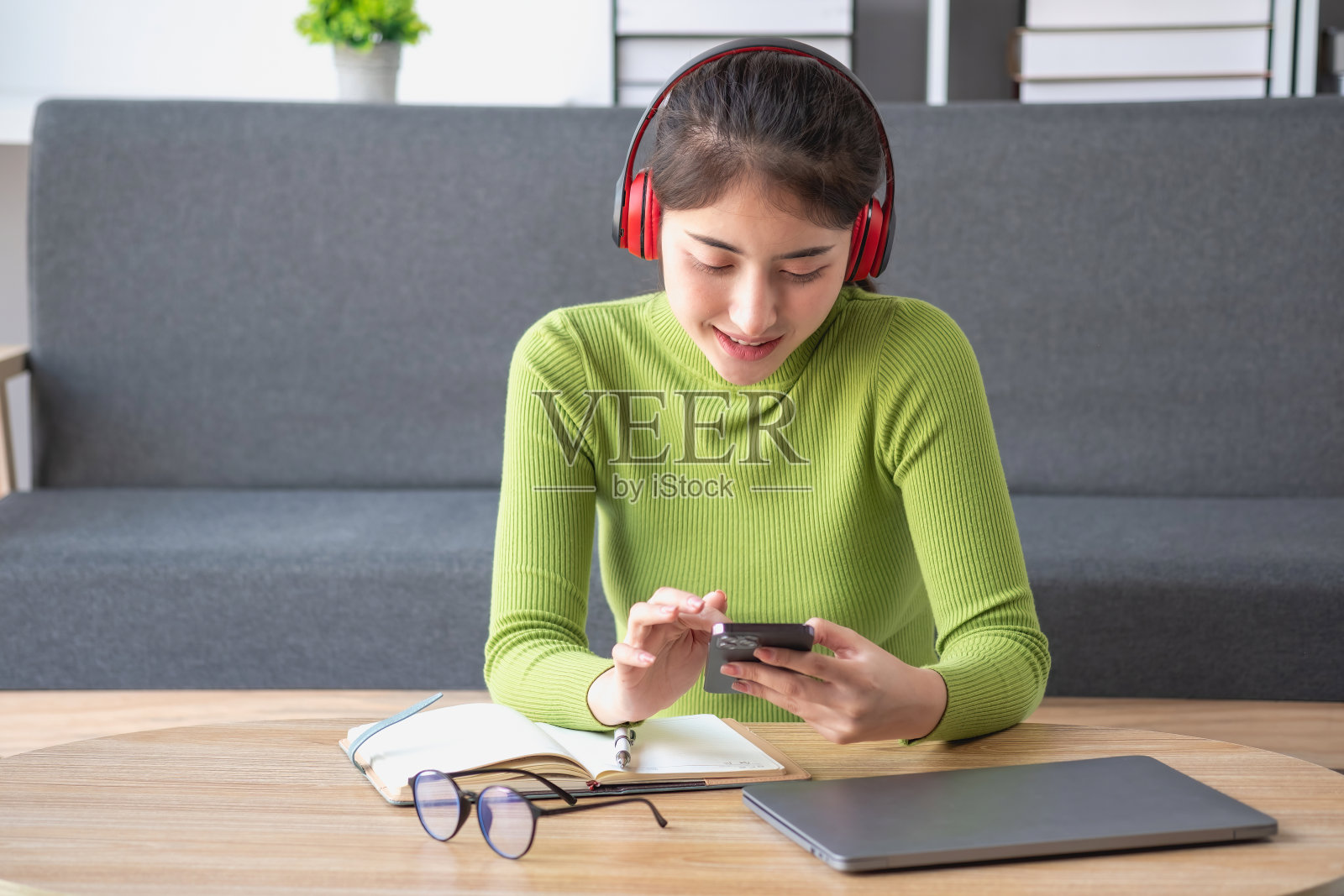 亚洲美女穿着绿色长袖衬衫，悠闲地坐在家里的客厅里，用笔记本电脑、手机和耳机看电影，听音乐。照片摄影图片