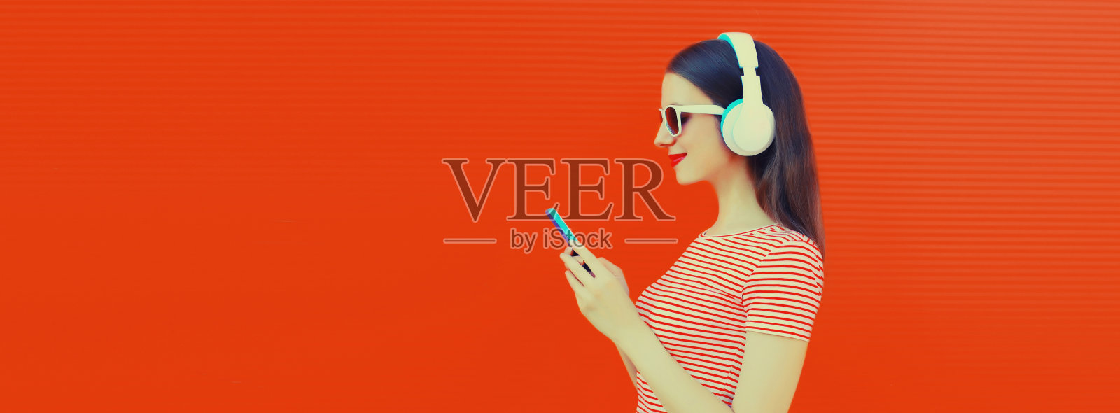 微笑的年轻女子戴着耳机，用橙色背景的智能手机听音乐照片摄影图片