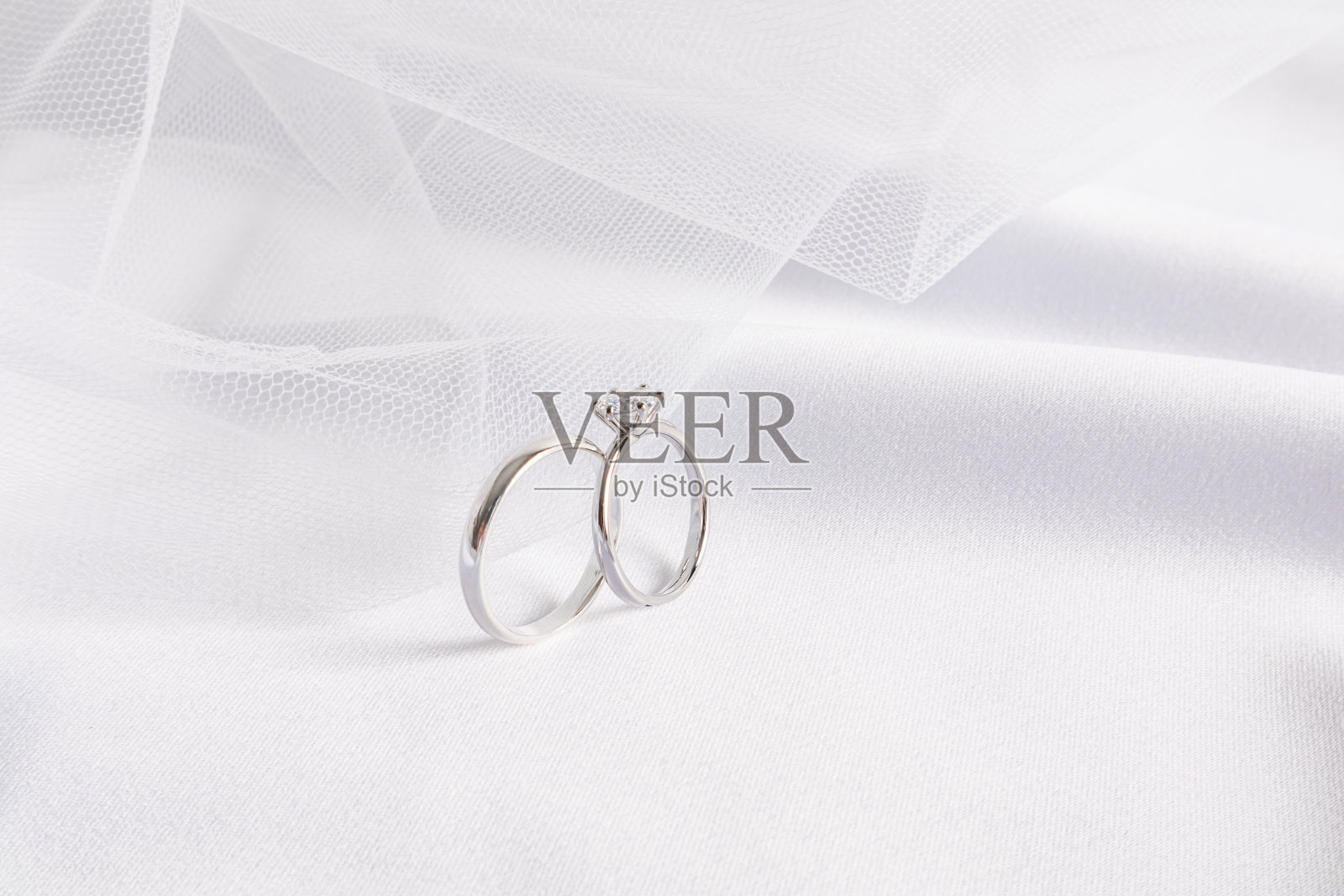 两个铂金和钻石订婚戒指的正面视图，背景是新娘的婚纱和柔软的缎面白色织物褶皱。照片摄影图片