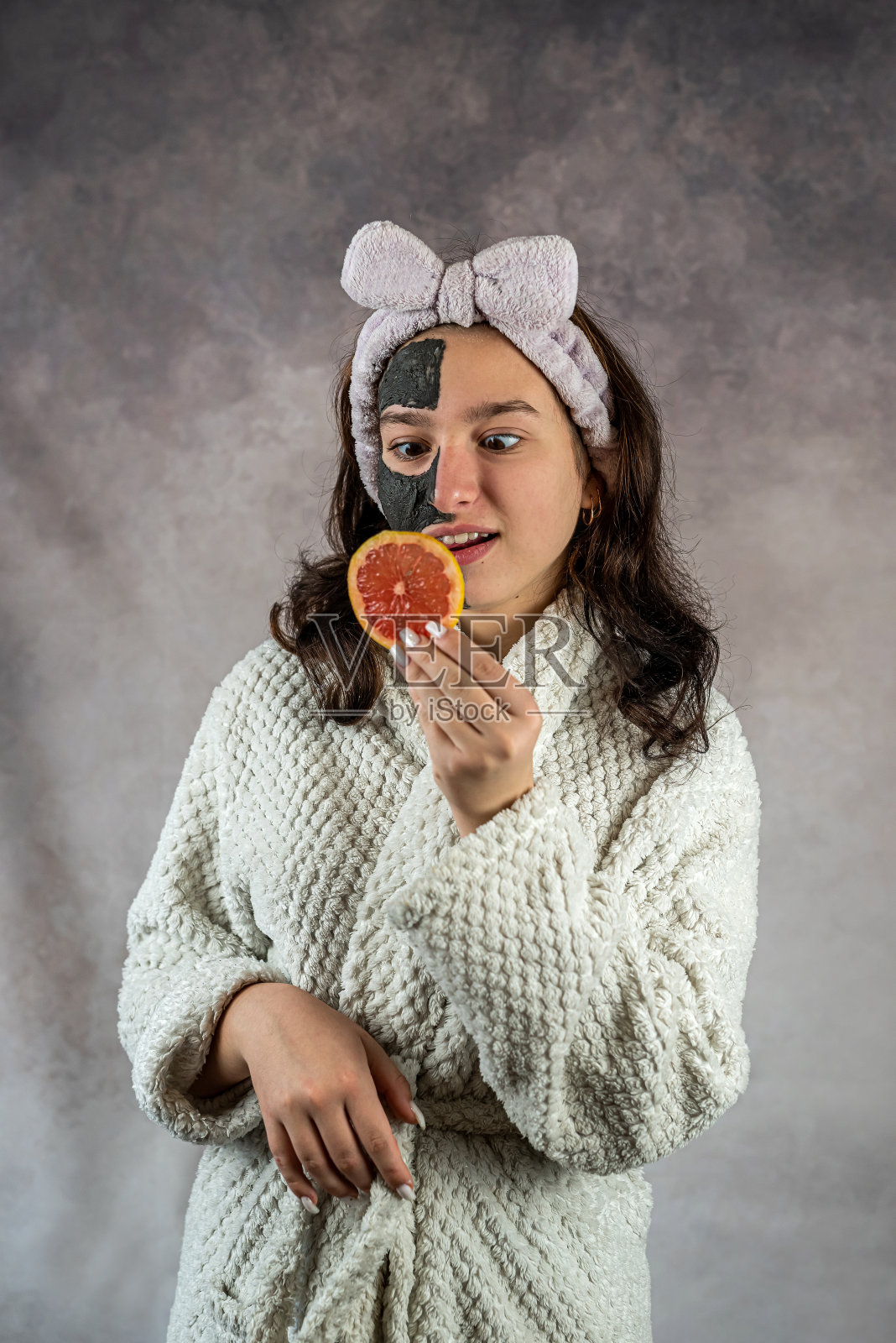 美丽的女孩穿着白色浴袍，戴着黑色的面膜，手里拿着柚子照片摄影图片