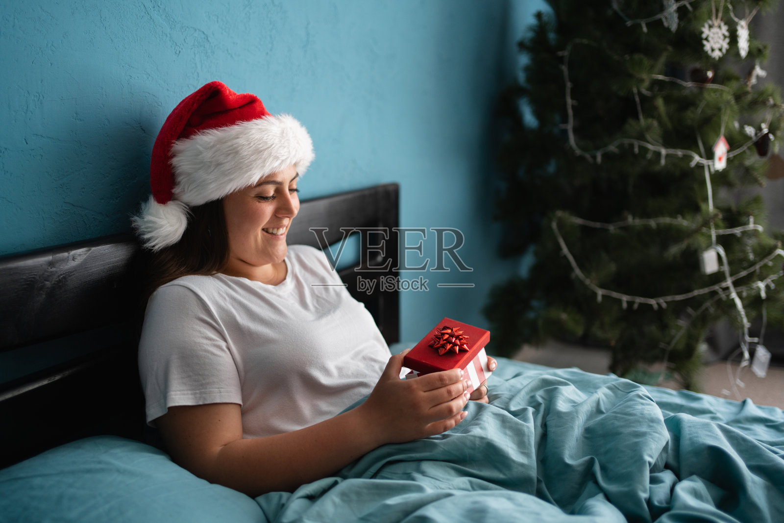 快乐的年轻女子在圣诞节早晨穿着睡衣在床上打开礼物照片摄影图片