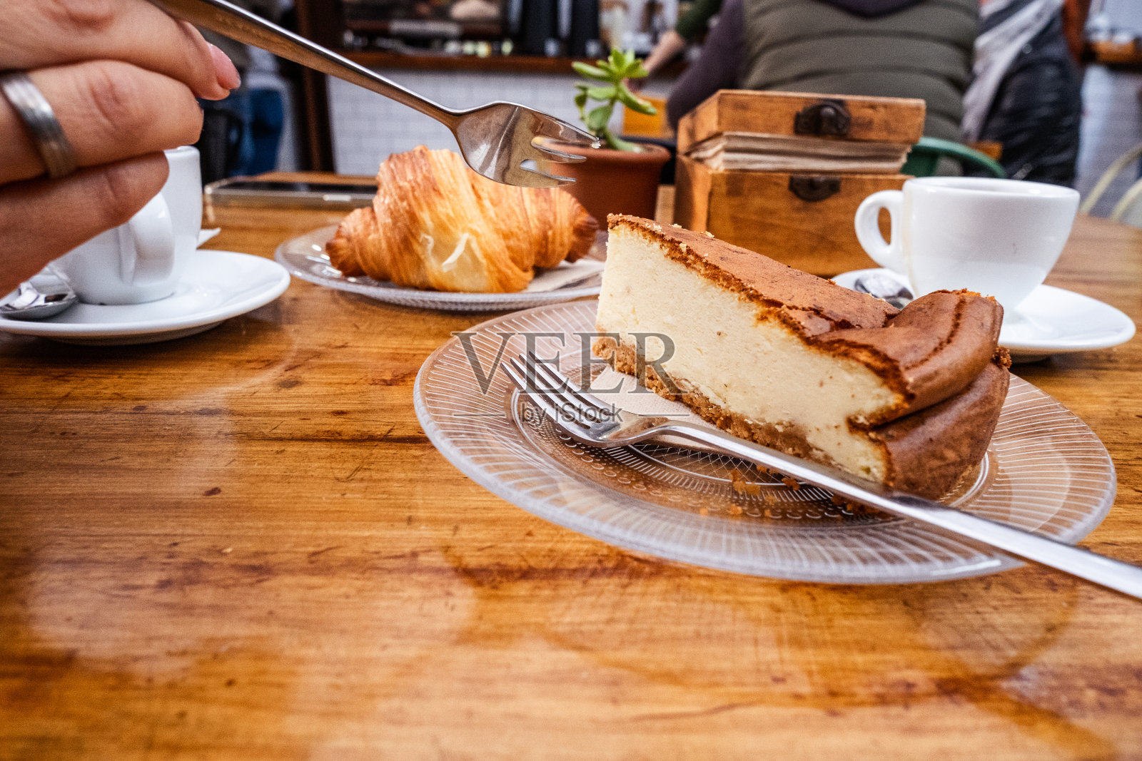 早餐。女人手里拿着叉子拿着一块芝士蛋糕放在一个玻璃板上，桌子上有一杯咖啡放在一个用金属勺子的盘子上，玻璃板上放着一个牛角面包和一杯茶。一切都在一张天然木桌上。美食。加那利群岛的特内里费岛照片摄影图片