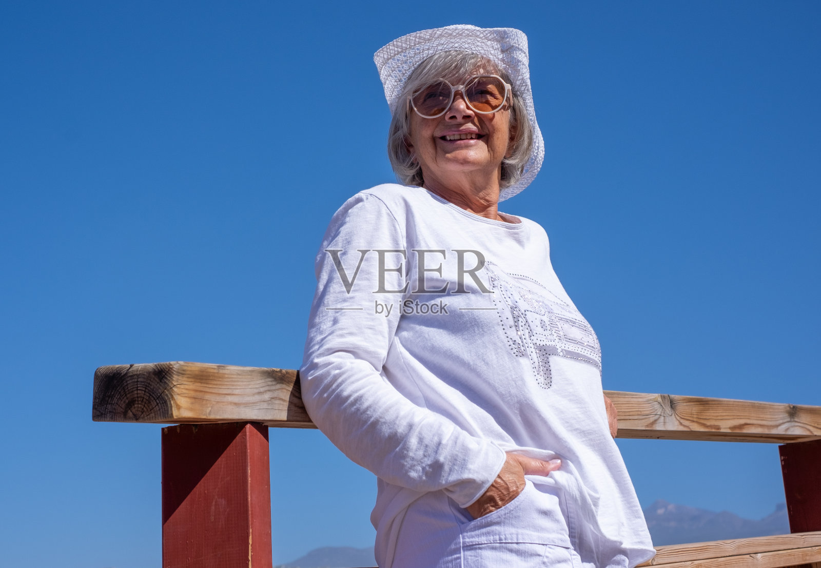在一个阳光明媚的日子里，戴着帽子、面带微笑的白衣老妇站在户外。享受自由和暑假的老太太照片摄影图片