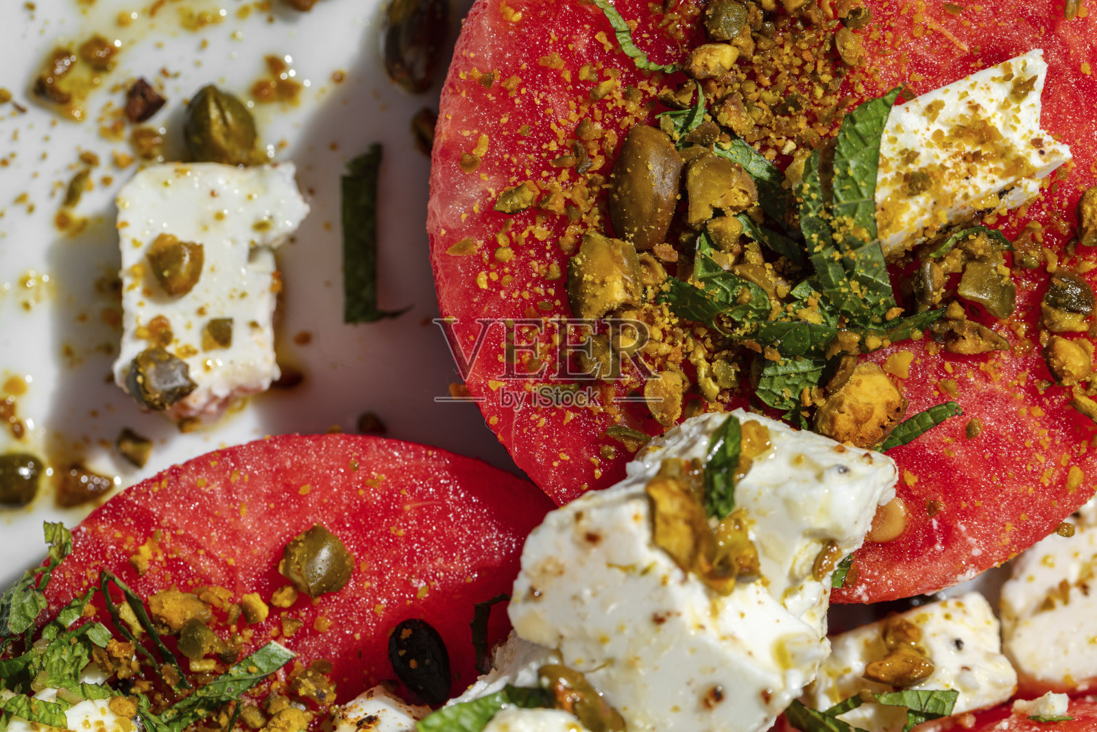 三片圆西瓜，配菲达奶酪、坚果和香草，淋上橄榄油。食物放在大理石台面上的一个轻陶瓷盘子里。旁边是一杯玫瑰酒和一瓶鲜花。照片摄影图片