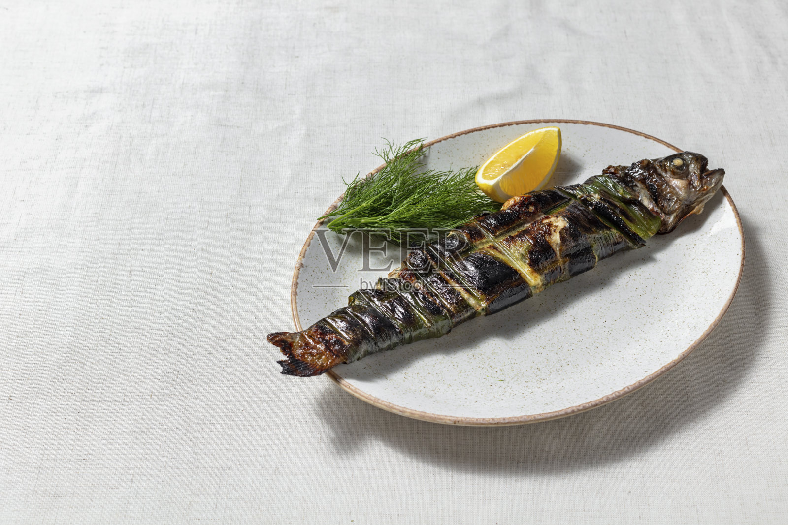 用葡萄叶、柠檬、斯万盐和香草烤金河鳟鱼。鳟鱼躺在一个椭圆形的、轻的陶瓷盘子里。旁边放着一片柠檬和一束莳萝。盘子放在一块轻薄的亚麻桌布上。照片摄影图片