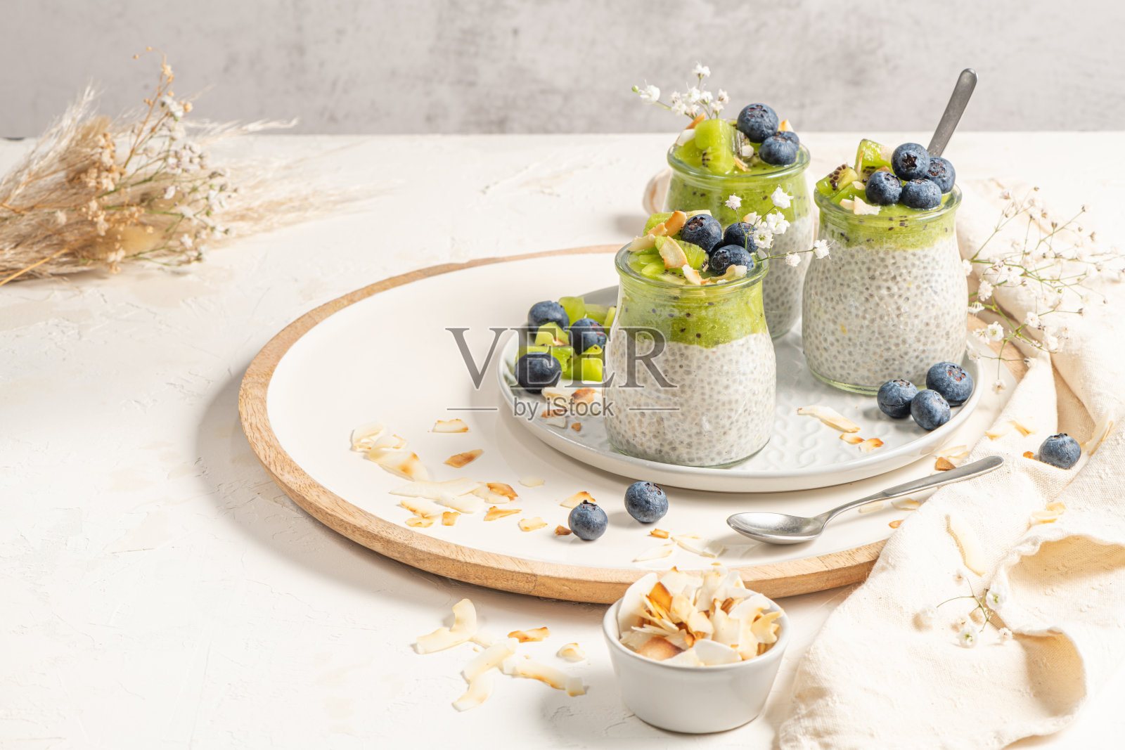 健康早餐奇亚布丁，有猕猴桃、蓝莓和椰子片，三份装在玻璃罐里，放在白色的桌子上。照片摄影图片