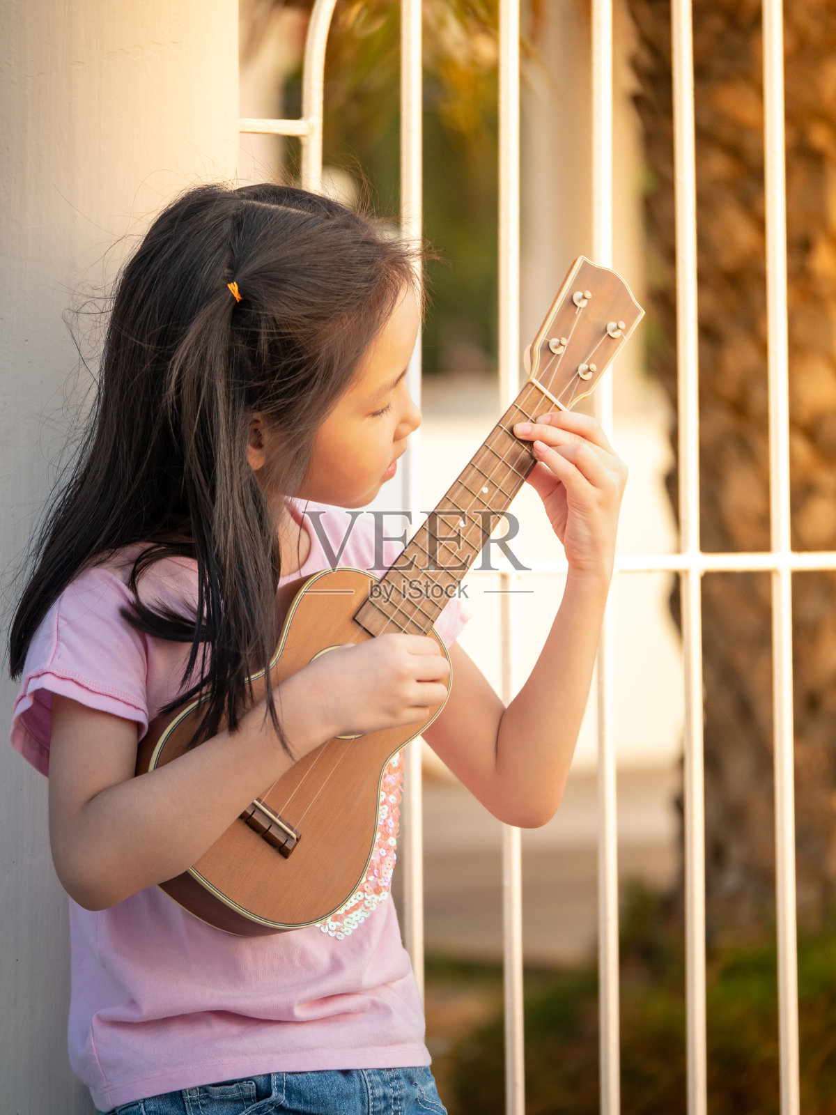 亚洲小女孩在花园里，倚着篱笆，在夕阳的余晖中练习弹奏尤克里里照片摄影图片
