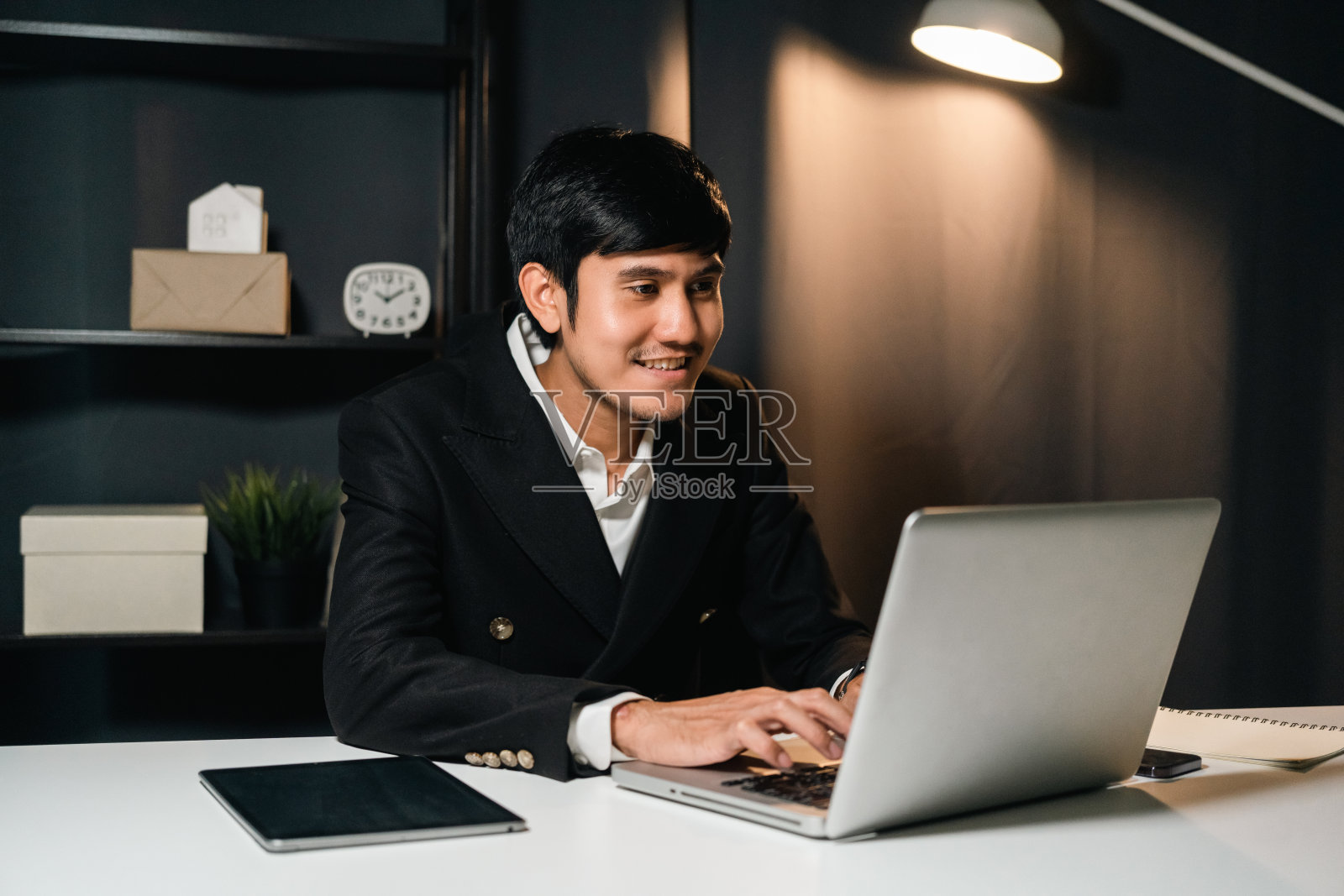 年轻的亚洲商人晚上在黑暗的办公室里用笔记本电脑工作。迷人英俊的印度男人在深夜带着落地灯的温暖灯光在家里努力加班照片摄影图片