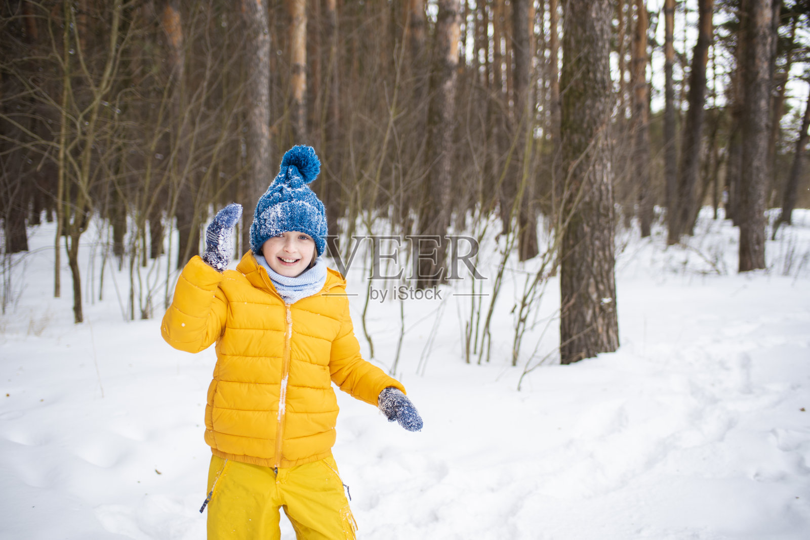 一个快乐微笑的孩子，穿着夹克，戴着帽子，围着围巾，在一个下雪的日子里走在冬天阳光明媚的森林里照片摄影图片