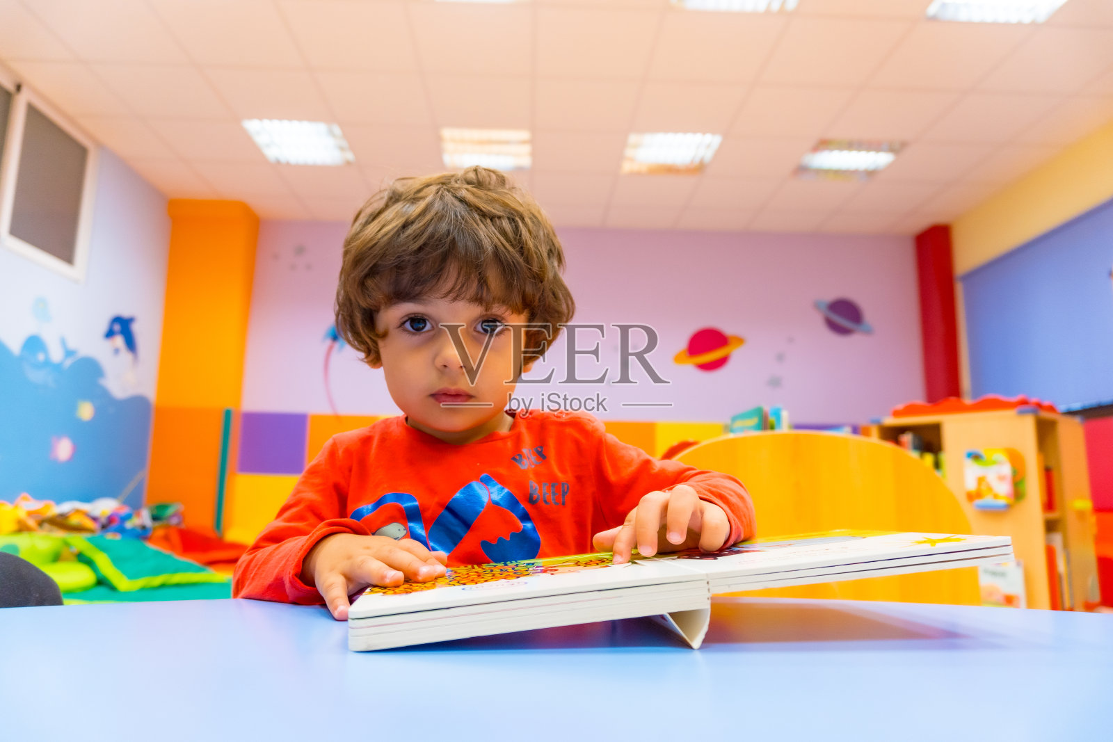 幼儿园室内，一个男孩坐在桌子旁看故事书，看着镜头照片摄影图片