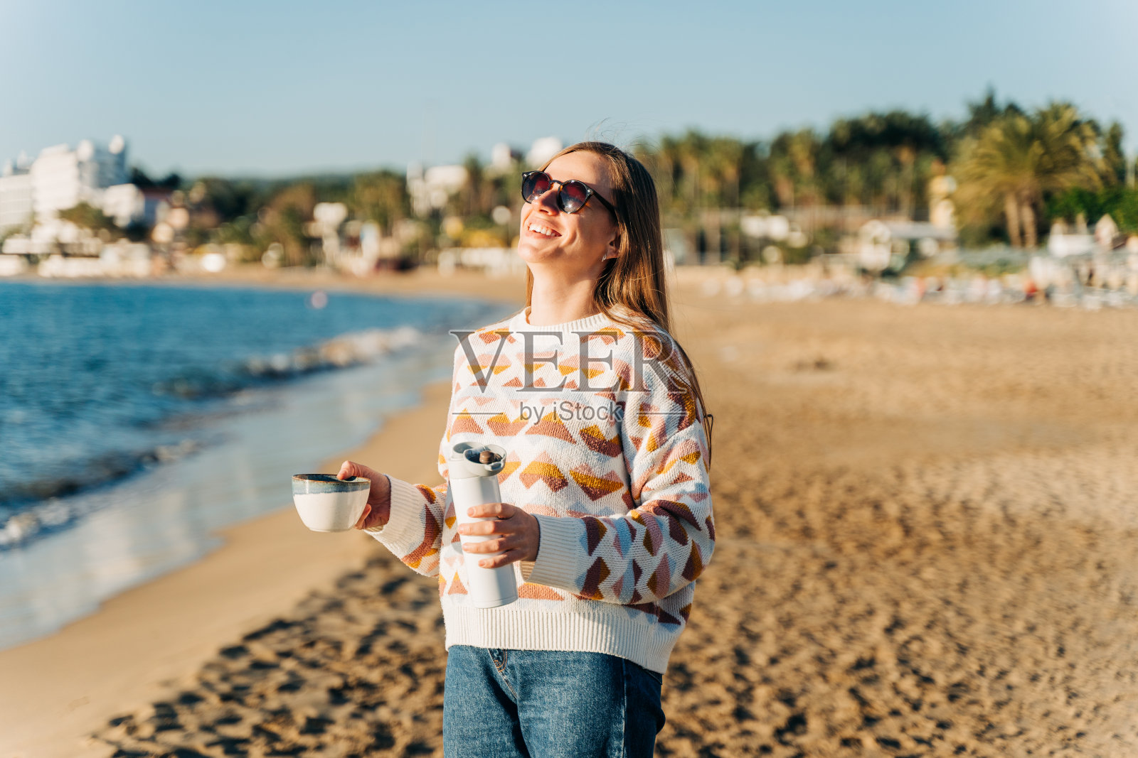 年轻美丽的女人在舒适的毛衣和太阳镜享受热咖啡从保温杯，而放松在冬天的海边沙滩。可爱迷人的女孩在海边喝茶晒太阳照片摄影图片