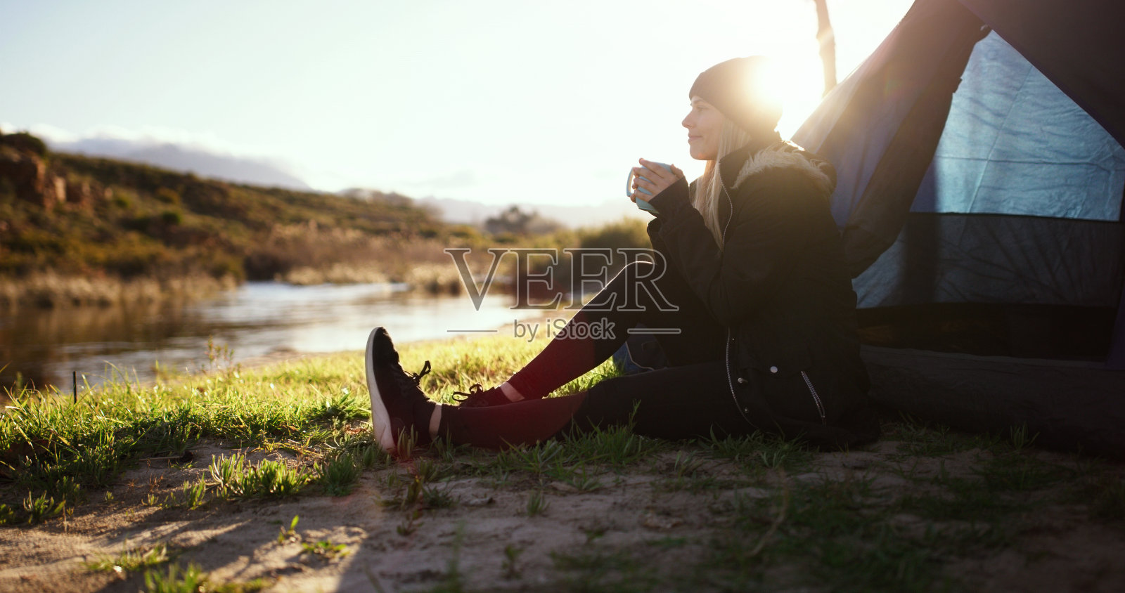 大自然，露营，女人端着一杯咖啡坐在帐篷边看日出。周末早晨在室外湖边喝着卡布奇诺的平静、放松的年轻女性。照片摄影图片