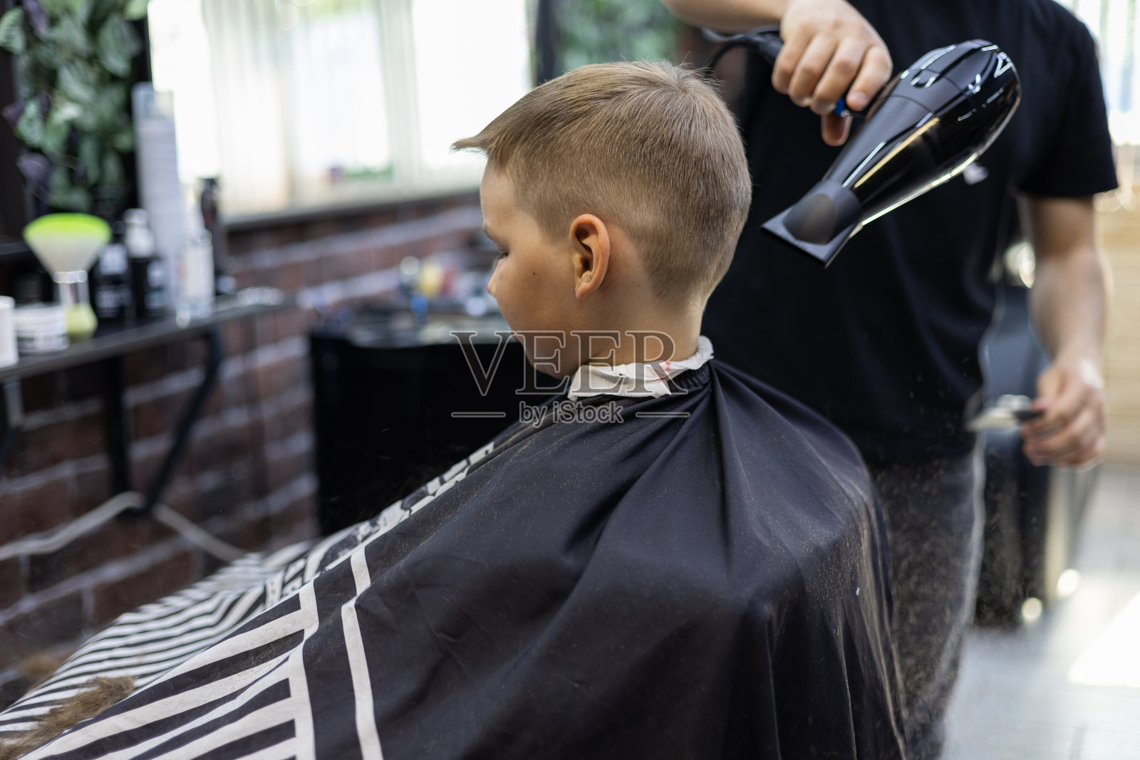 在吹风机的帮助下，公园管理员吹掉了男孩孩子脖子和肩膀上剪下来的头发。理发师为这个夏天的孩子做了一个时髦的发型。照片摄影图片