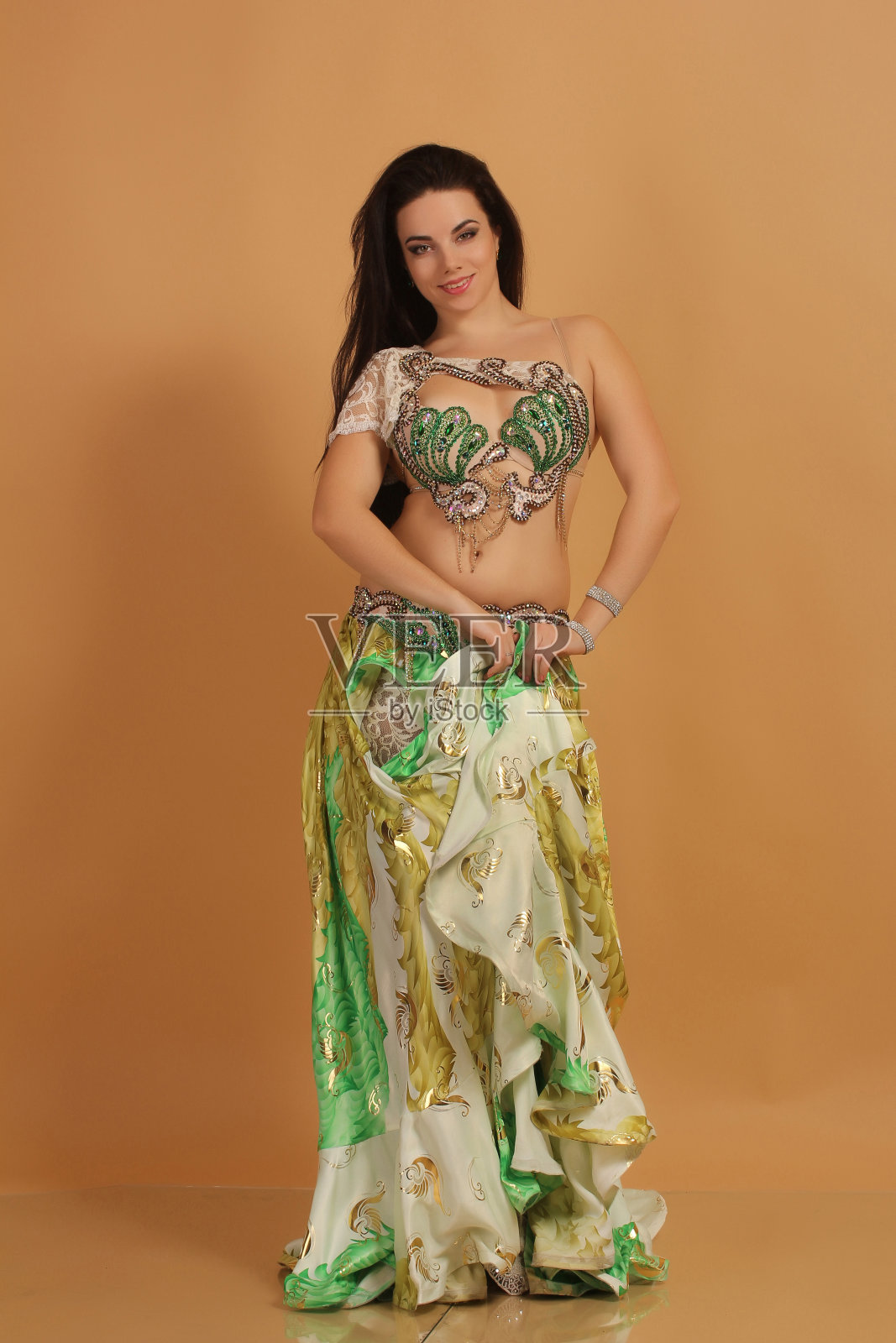 一个漂亮的黑发女孩跳着go-go和肚皮舞，在工作室里穿着绿色的阿拉伯连衣裙，在米色的背景上伸展姿势照片摄影图片