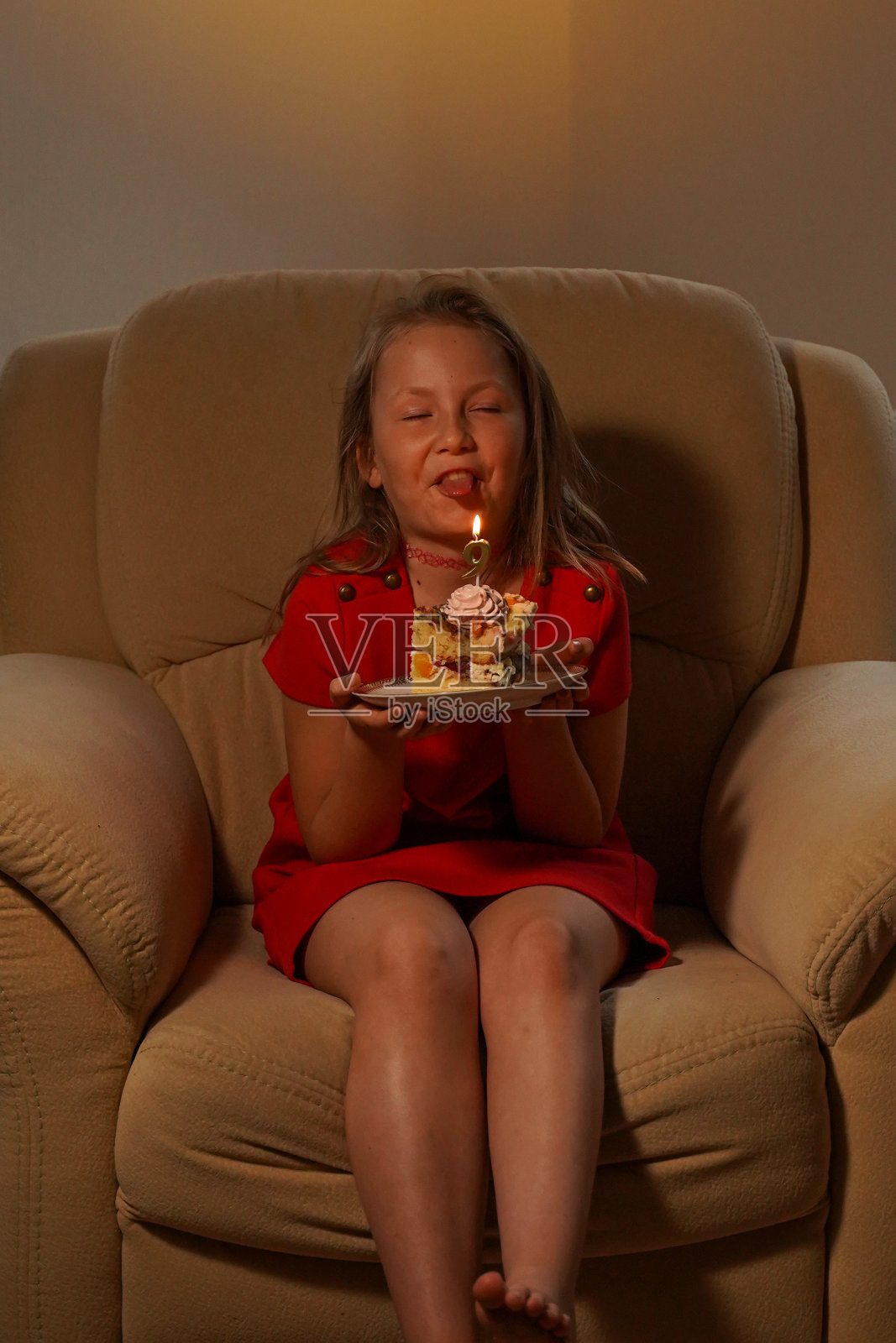 一个女孩坐在扶手椅上，手里拿着一个生日蛋糕，做着鬼脸。生日快乐。照片摄影图片